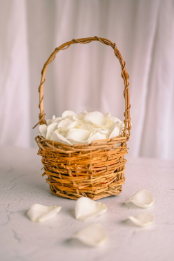 A small wicker basket filled with white rose petals on a white surface with scattered petals beside it.