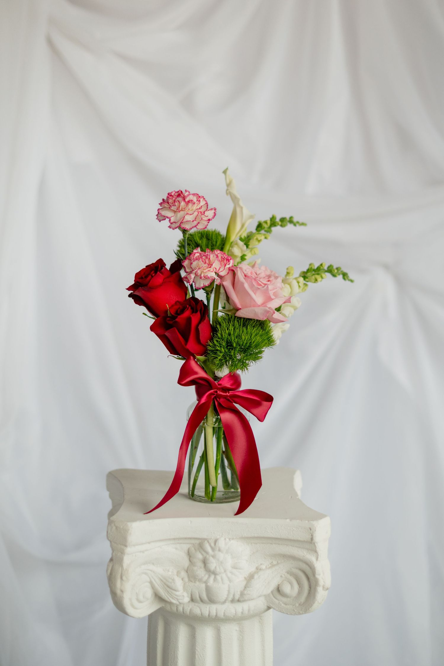 Elegant floral arrangement with red and pink roses in a glass vase adorned with a red bow sits atop a white decorative pedestal.