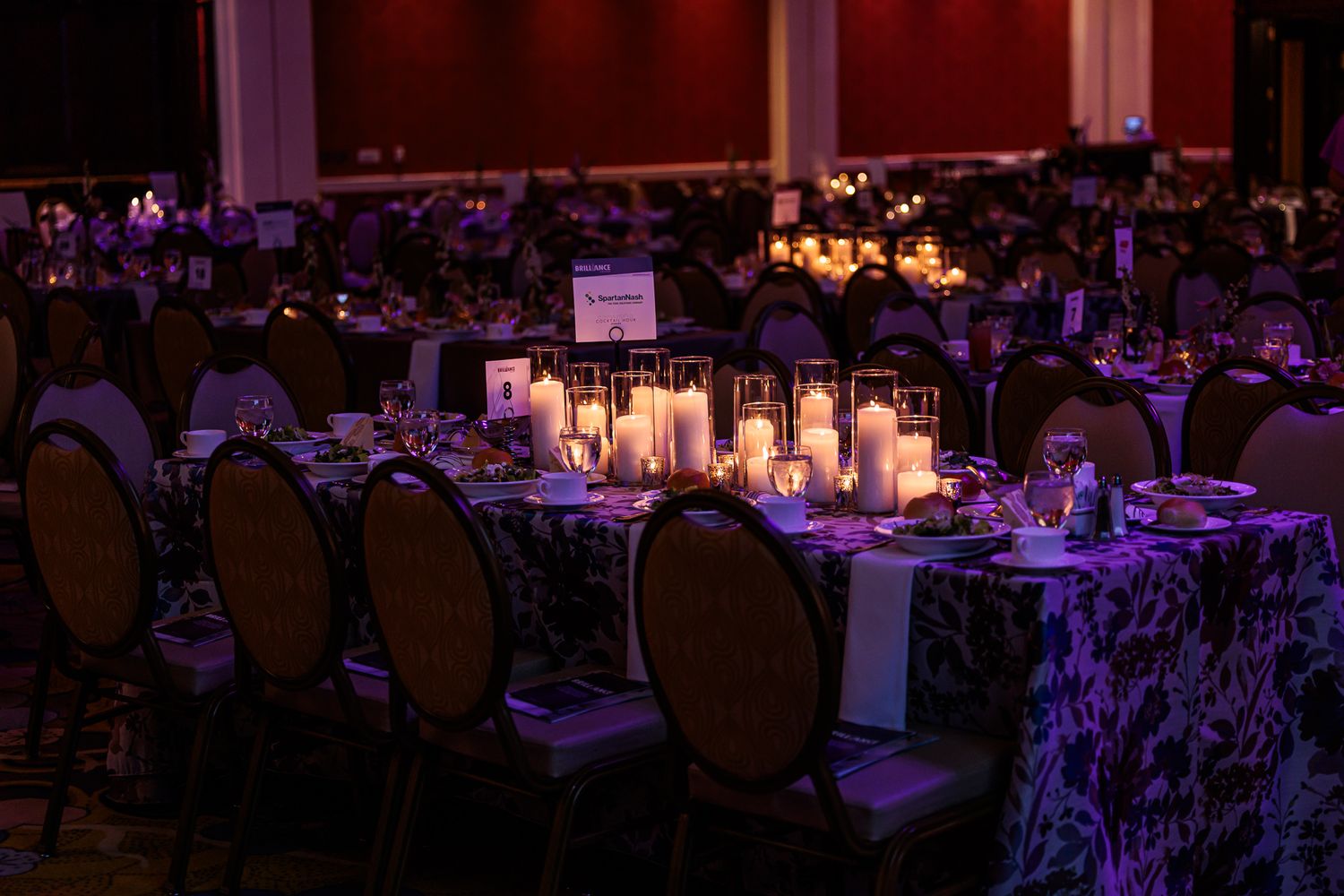 Elegant banquet table setting with glowing candles and dark upholstered chairs in a dimly lit ballroom.