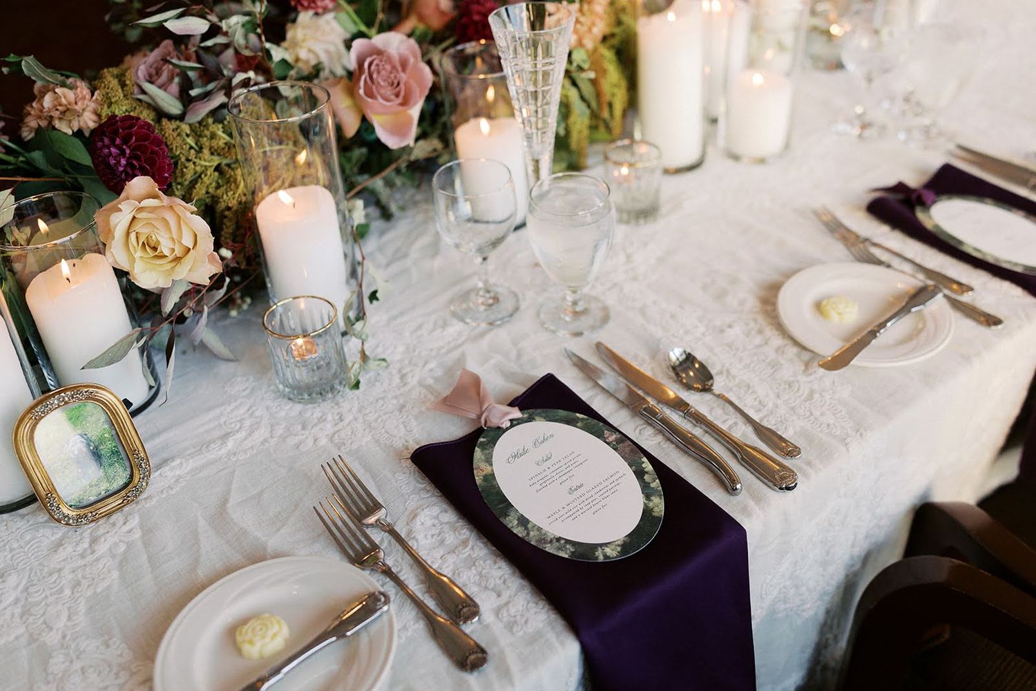 Elegant wedding reception table setting with white lace tablecloth, purple napkins, candles, and floral centerpiece.