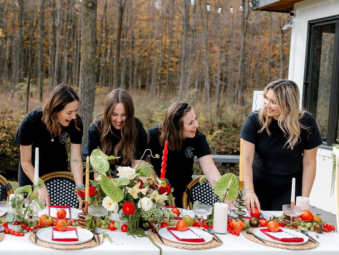 Four women in black shirts arrange a festive outdoor tablescape with florals, red accents, and autumn trees in the background.