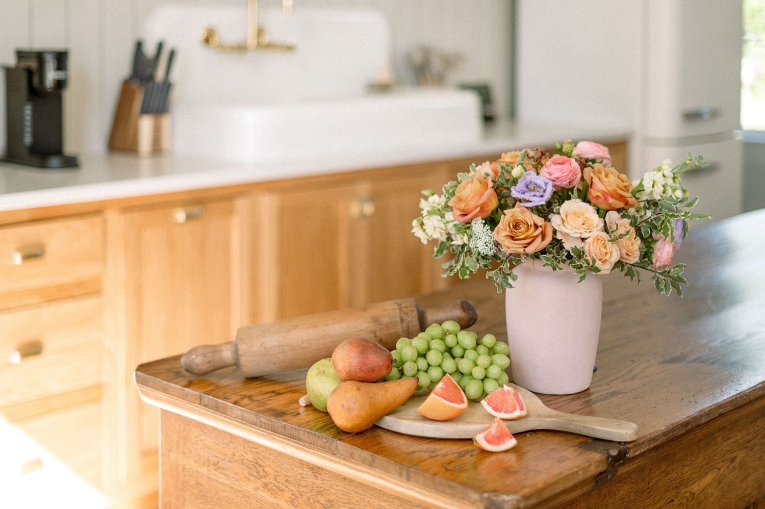 Fresh floral arrangement in pink vase with fruit and snacks on wooden counter in bright modern kitchen.