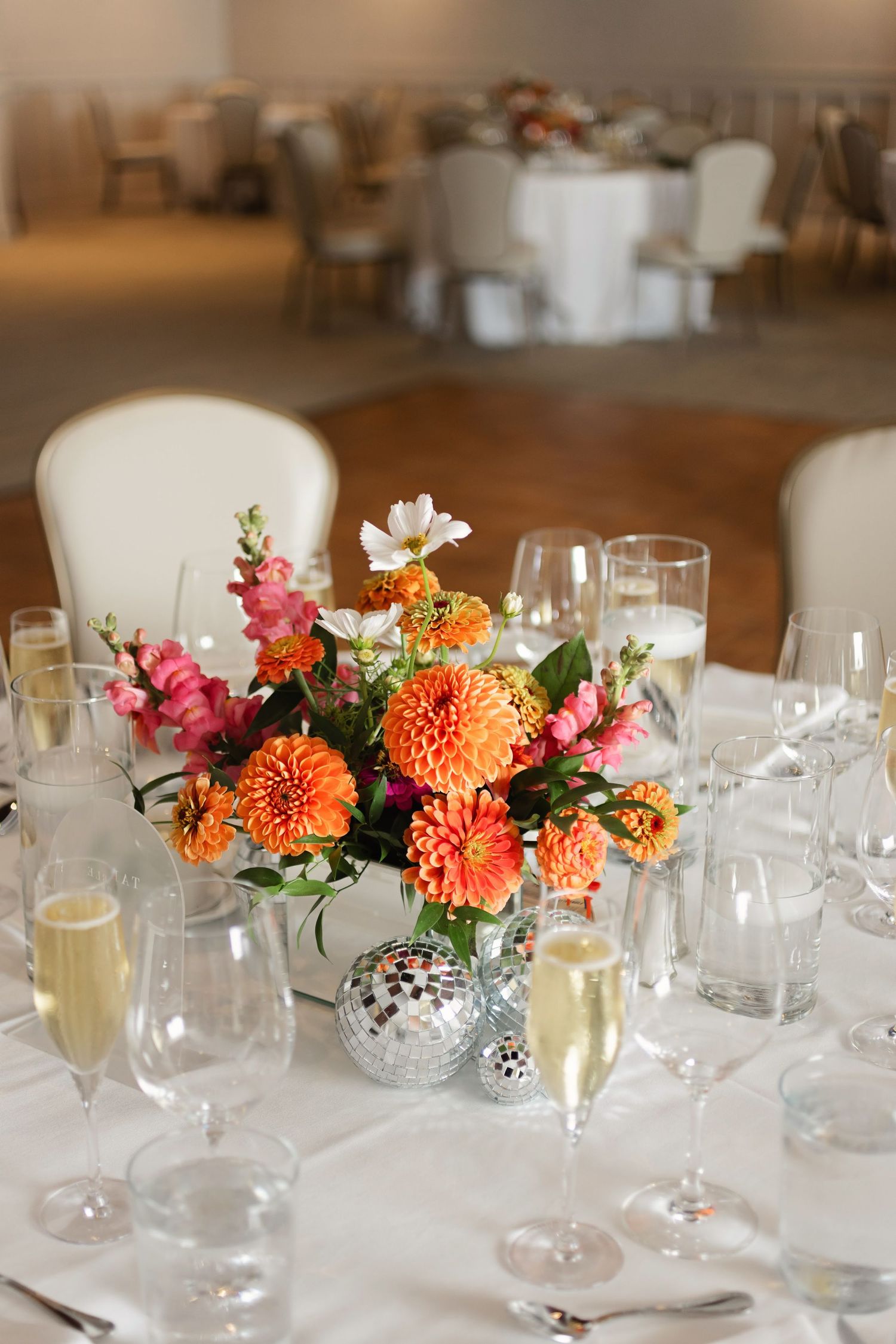 Elegant wedding table setting with orange dahlia and pink flower centerpiece alongside silver disco balls and champagne glasses.