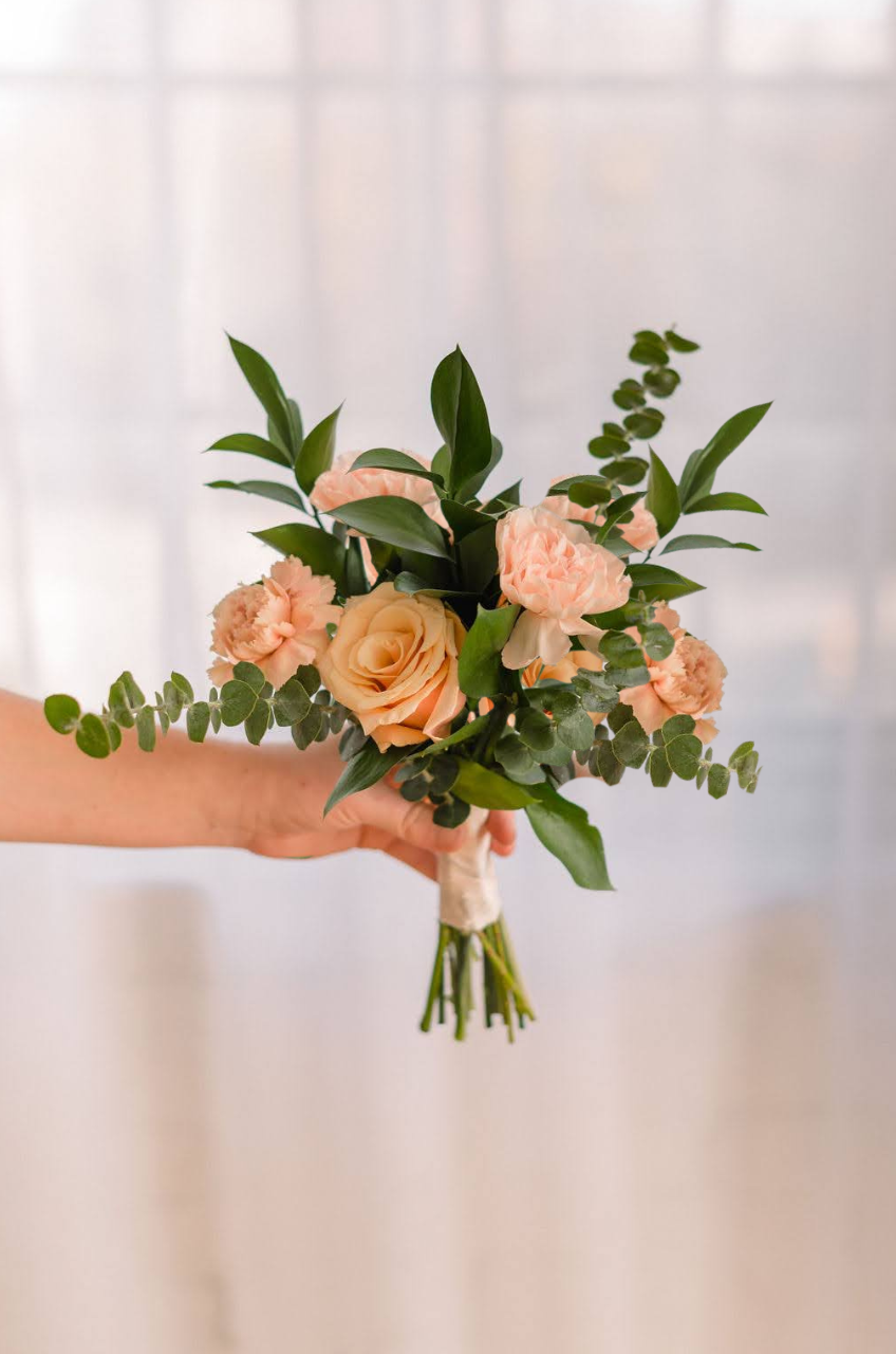 Hand holding a small bridal bouquet of peach roses, carnations, and eucalyptus greenery against a soft white background.