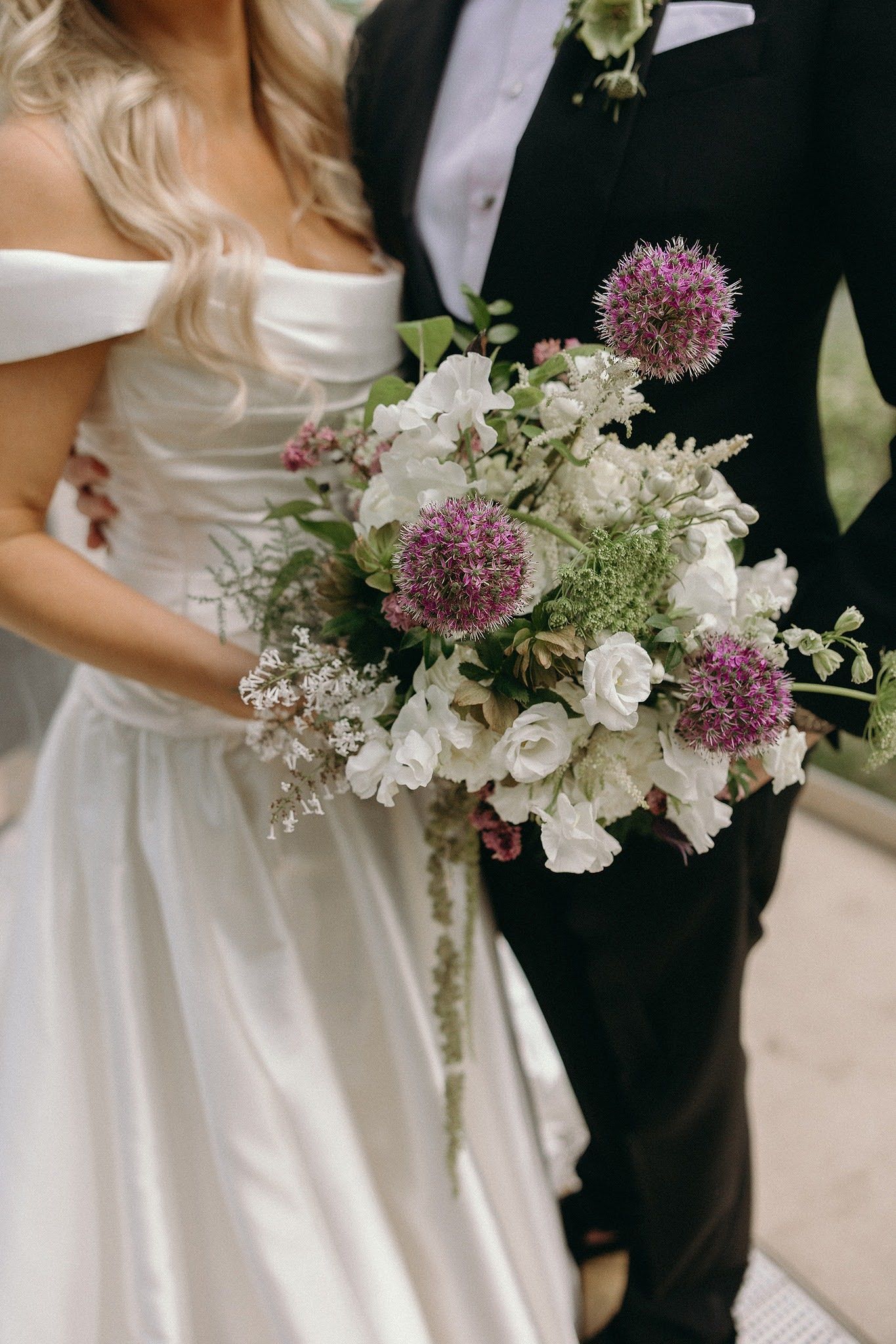 Bride and groom holding a lush wedding bouquet of white roses and purple allium flowers.