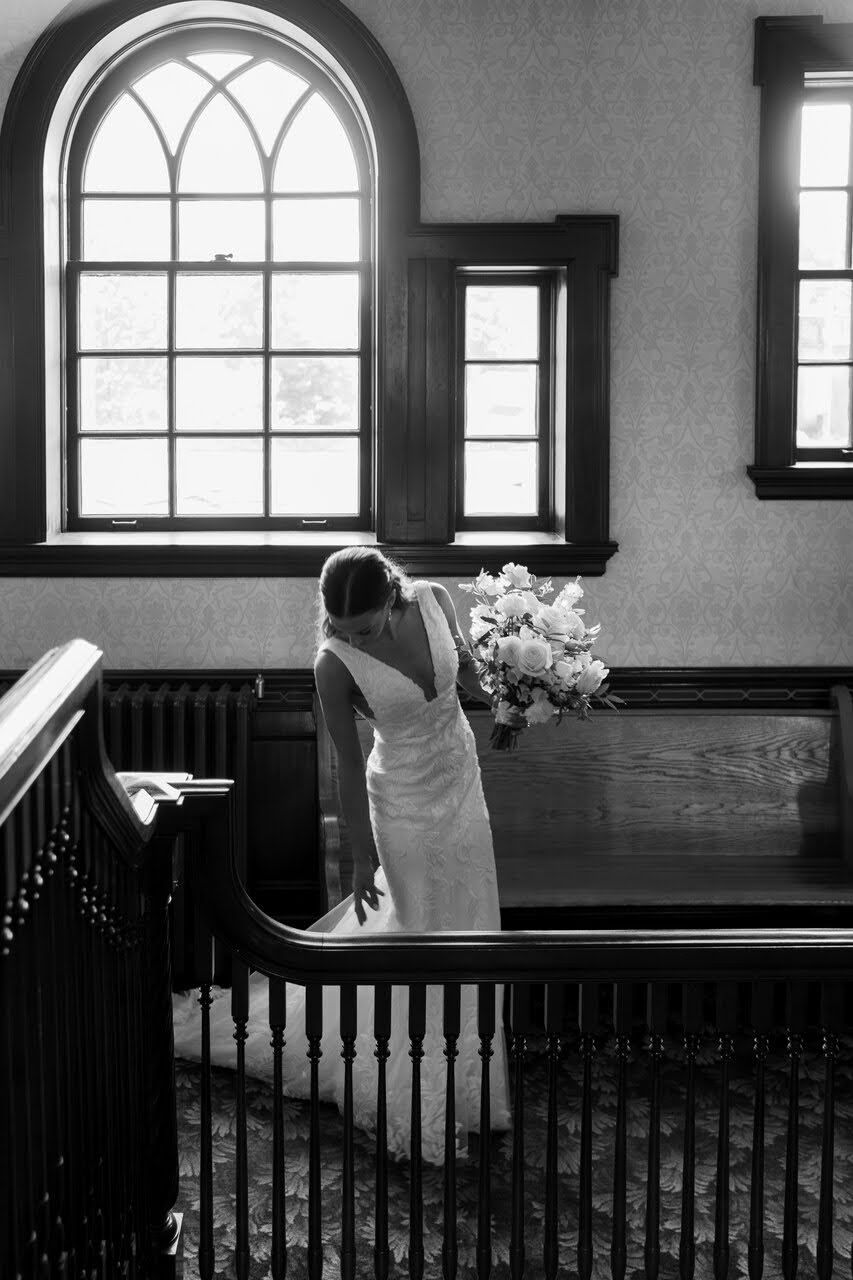 Black and white photo of a bride in a white gown holding a bouquet inside a church with arched windows.