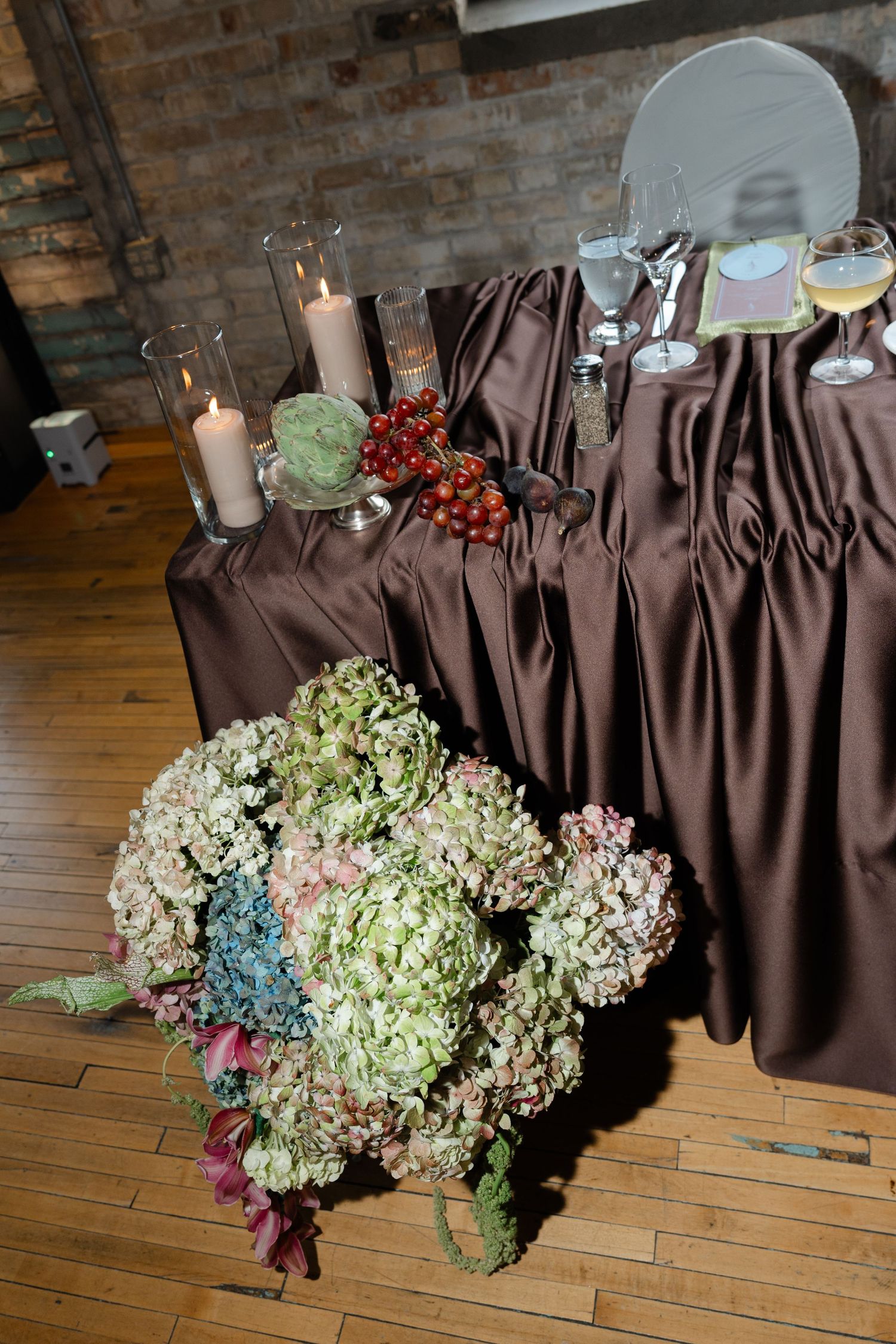 Elegant wedding reception table with brown satin cloth, hydrangeas, candles, red berries, and wine glasses on hardwood floor.