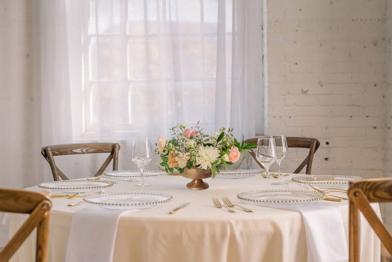 Elegant wedding table setting with blush floral centerpiece, crystal glasses, gold cutlery, and ivory linen in a bright white room.