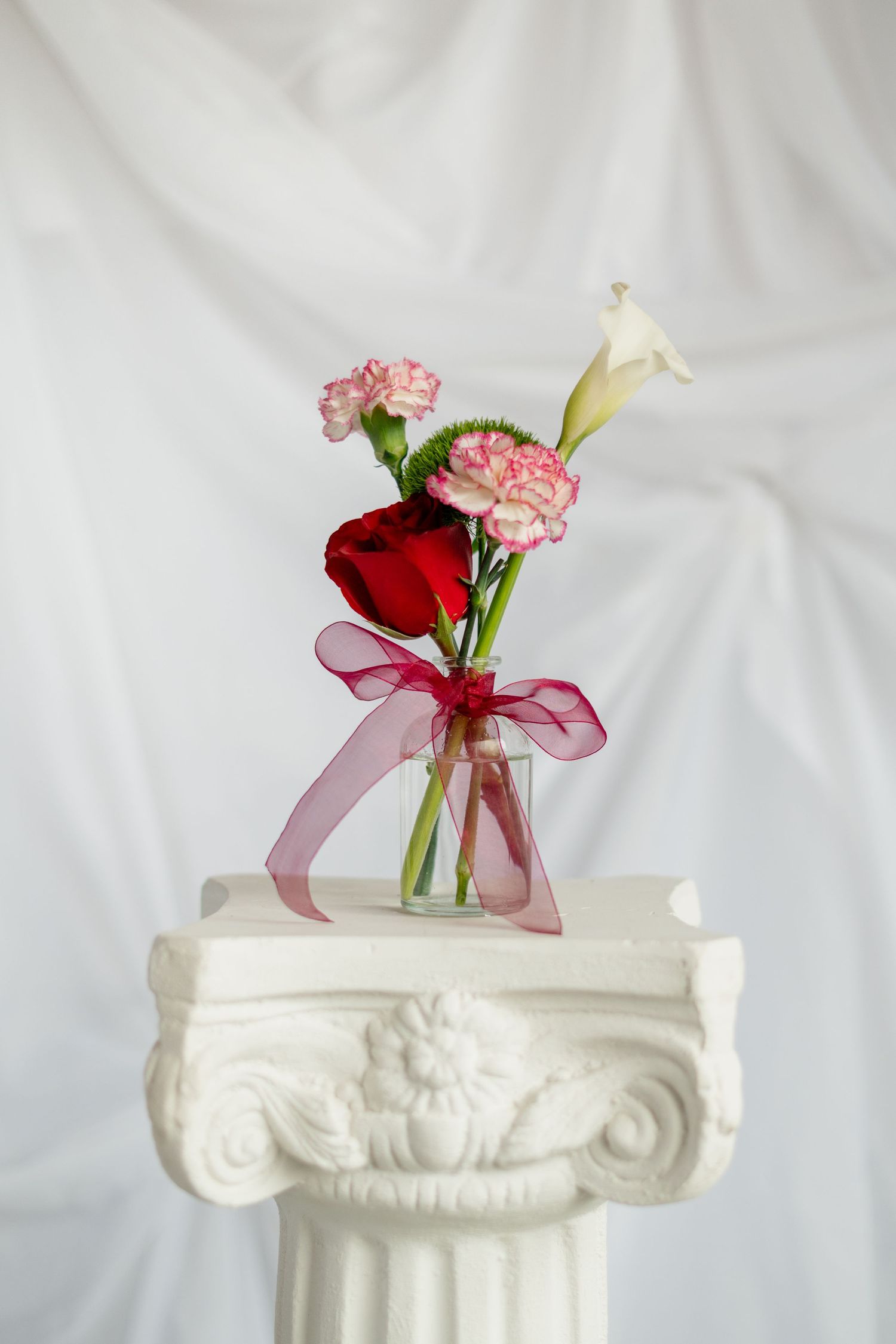 White decorative column pedestal with red and pink carnations in small glass vases tied with burgundy ribbon.