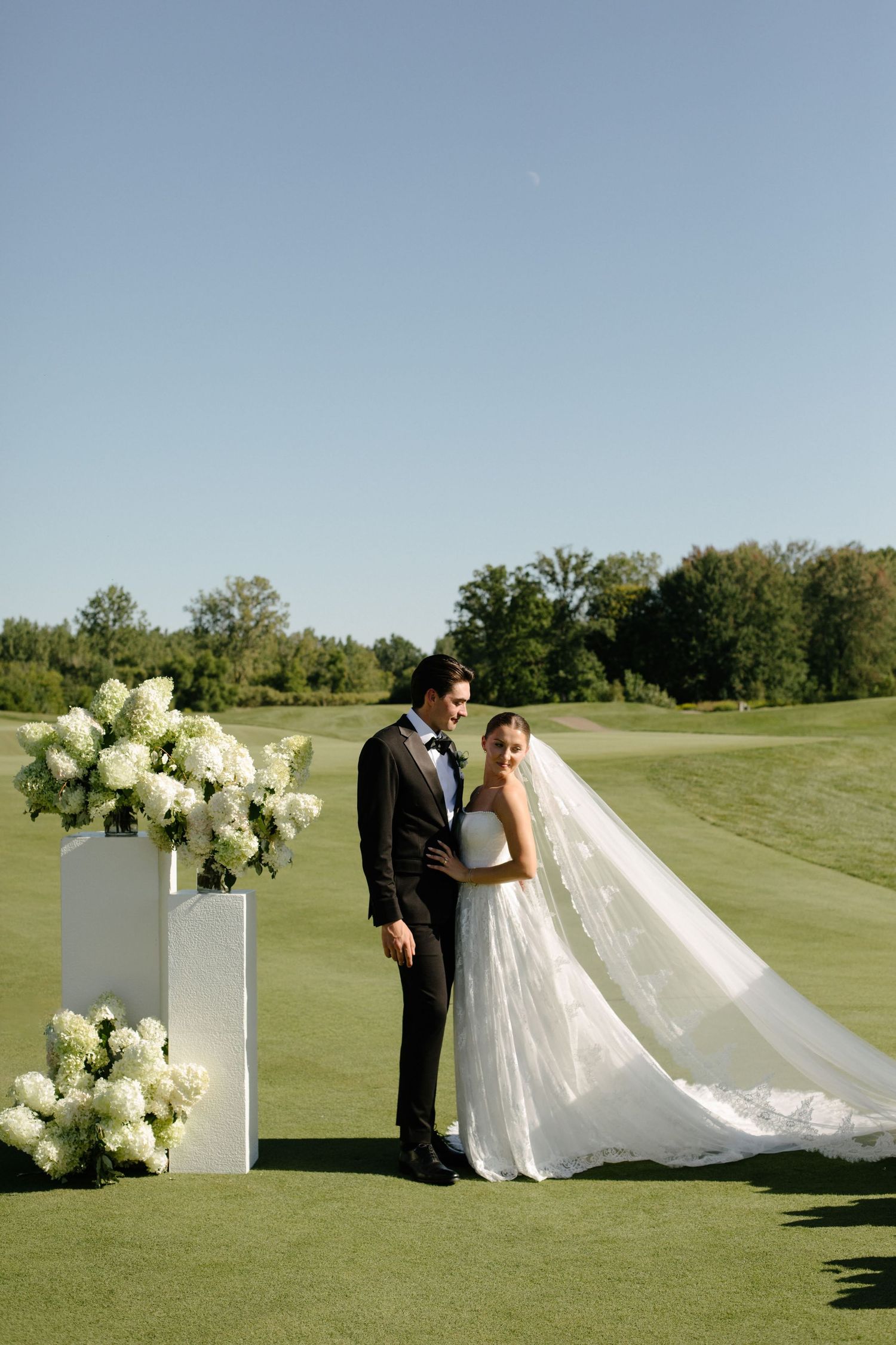 Bride and groom stand together on lush green golf course beside white floral pedestals with hydrangeas under clear blue sky.