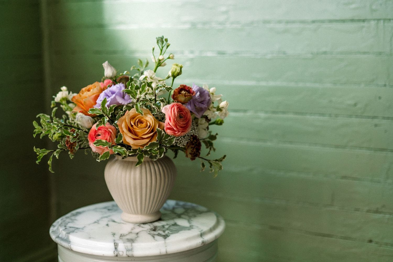 A colorful floral arrangement with orange roses and mixed blooms in a white ribbed vase sits on a marble table against green wall.