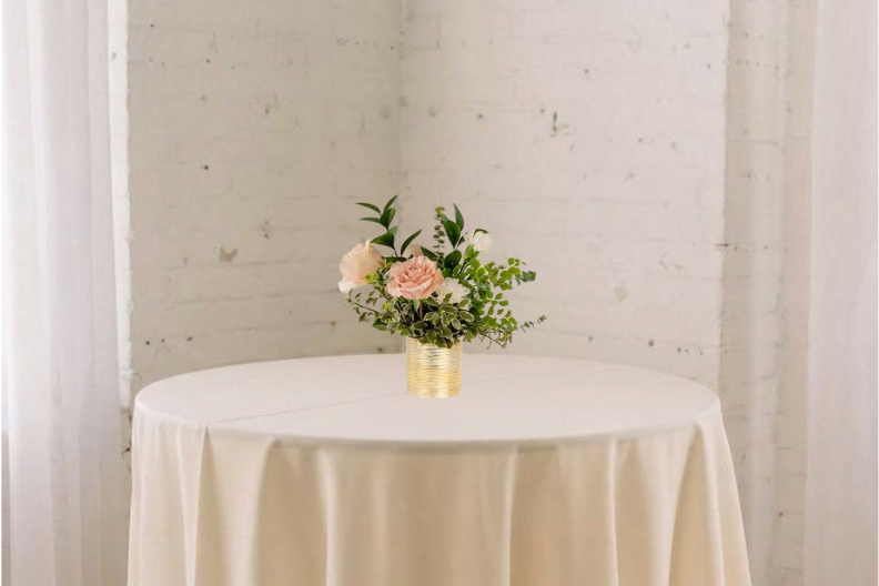 Round table with cream linen and a small pink floral centerpiece in a gold vase against a white brick wall.