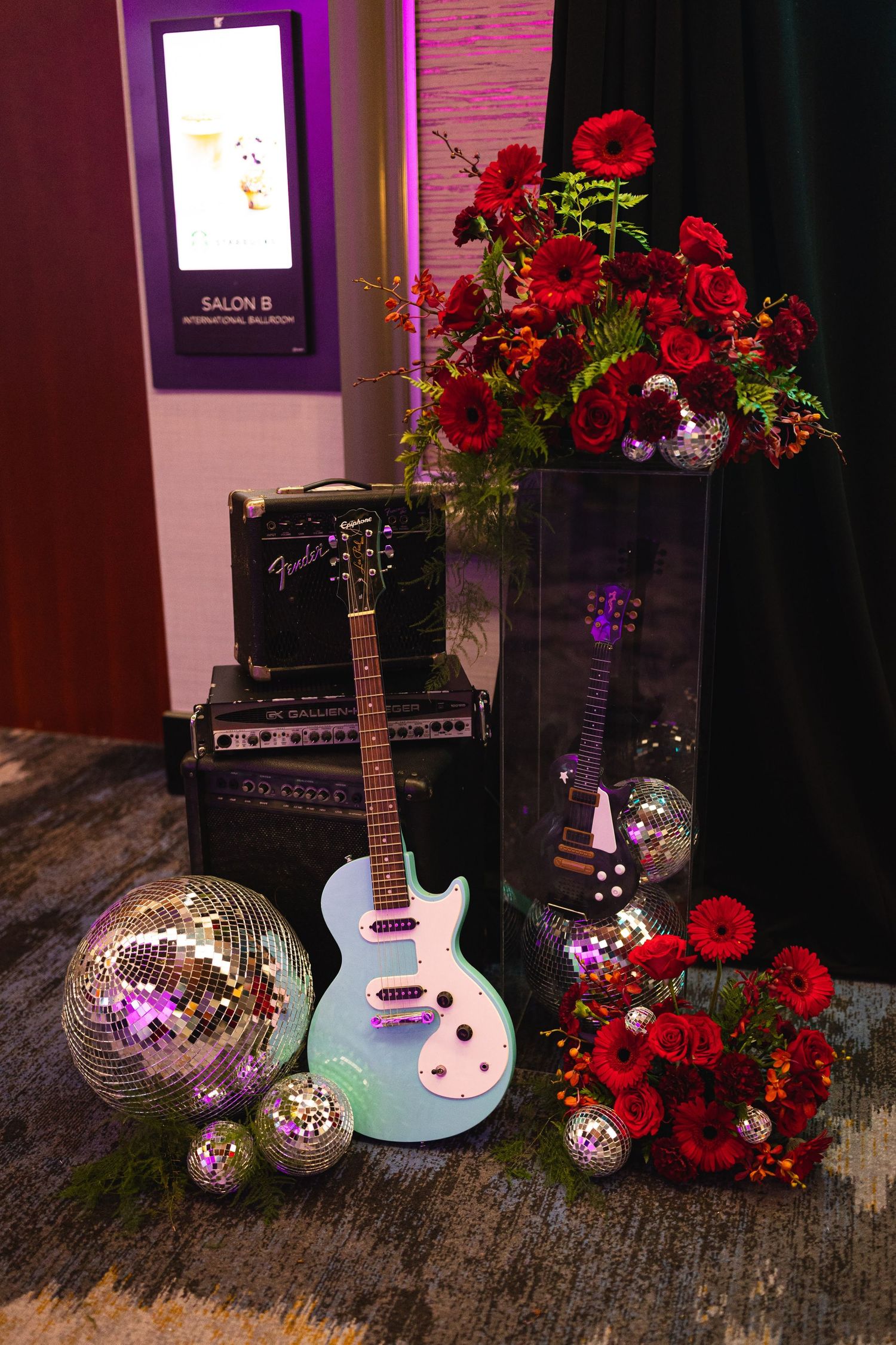 A light blue electric guitar sits beside disco balls and red roses on a rustic wooden surface in a dimly lit setting.