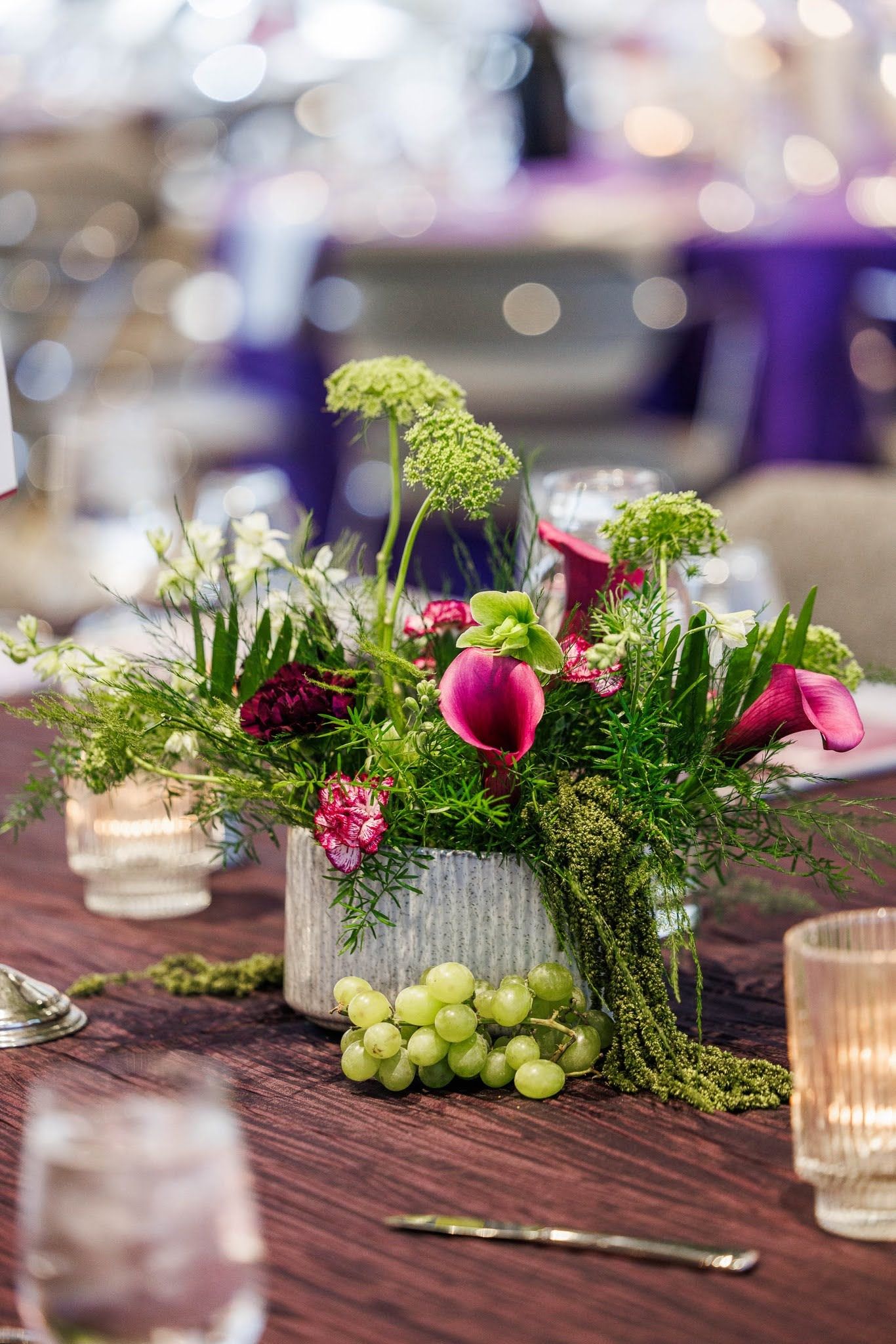 Elegant floral centerpiece with pink calla lilies, green blooms, and fresh grapes on a rustic wooden table at a formal event.