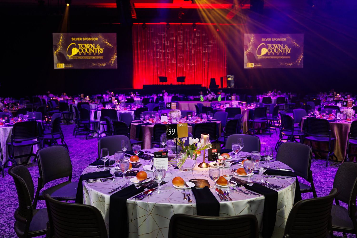 Elegant banquet hall setup with round tables and dramatic purple and red lighting creating an atmospheric ambiance.