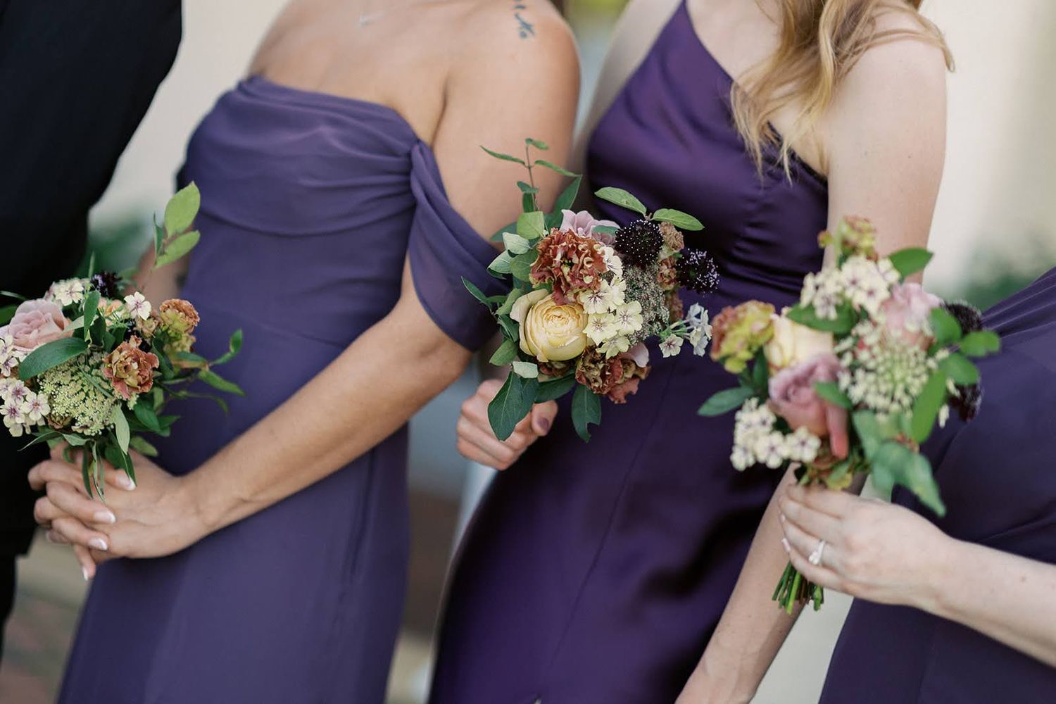 Bridesmaids in purple satin gowns holding rustic bouquets with pink, yellow, and greenery at a wedding.
