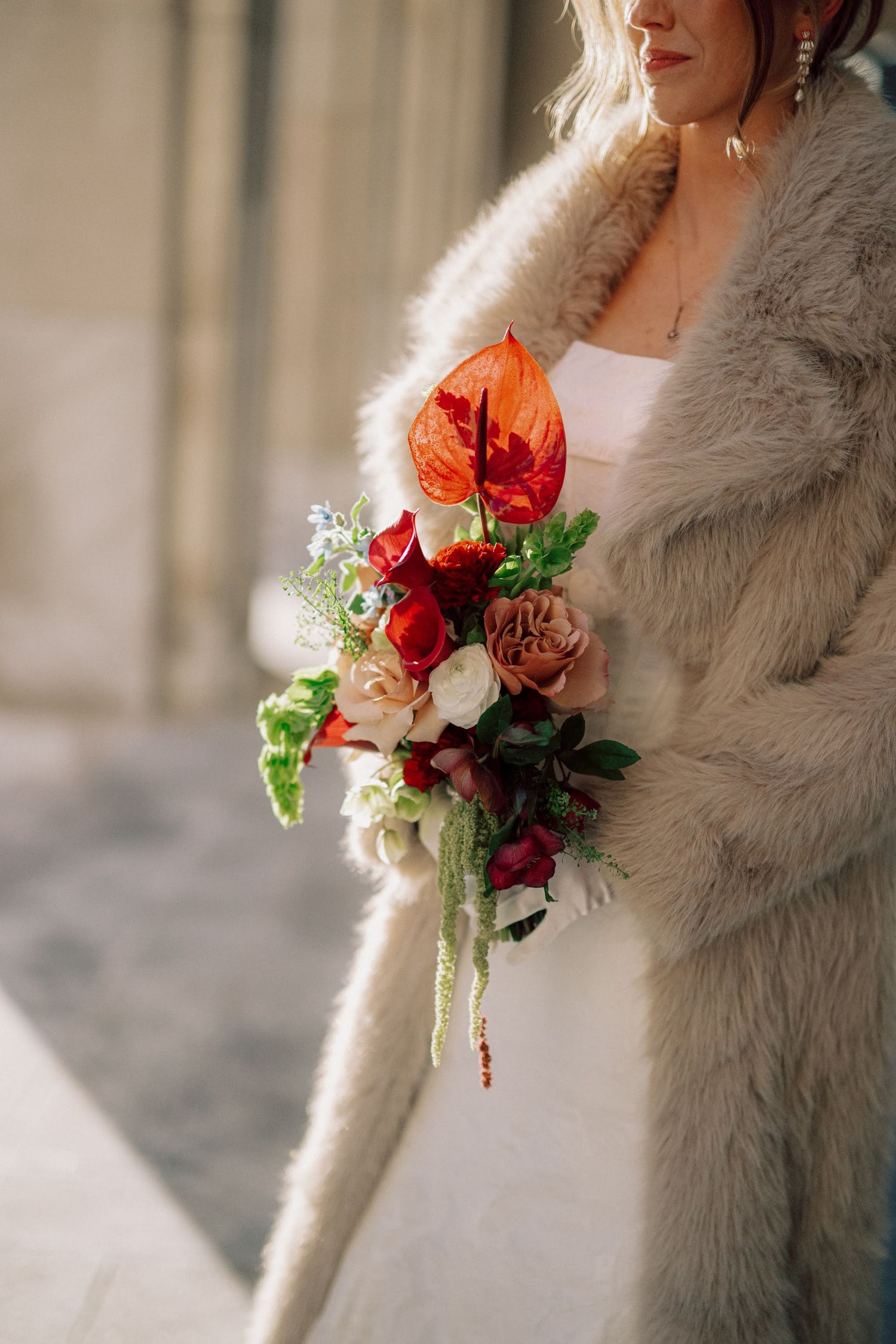 A bridal bouquet with red and white flowers is held against a soft grey fur coat in an elegant wedding photo.