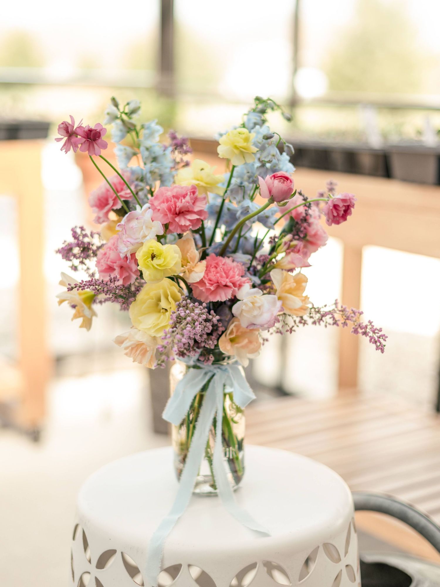 A delicate floral arrangement with pink carnations, yellow daffodils, and blue delphinium in a glass vase with ribbon accent.