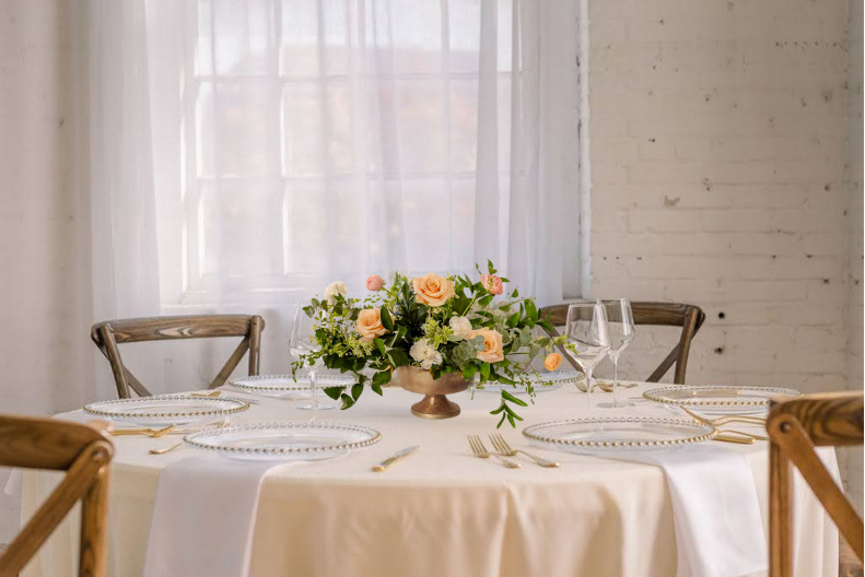 Elegant wedding table setting with peach floral centerpiece, gold accents, crystal glasses, and neutral linen in a bright white room.