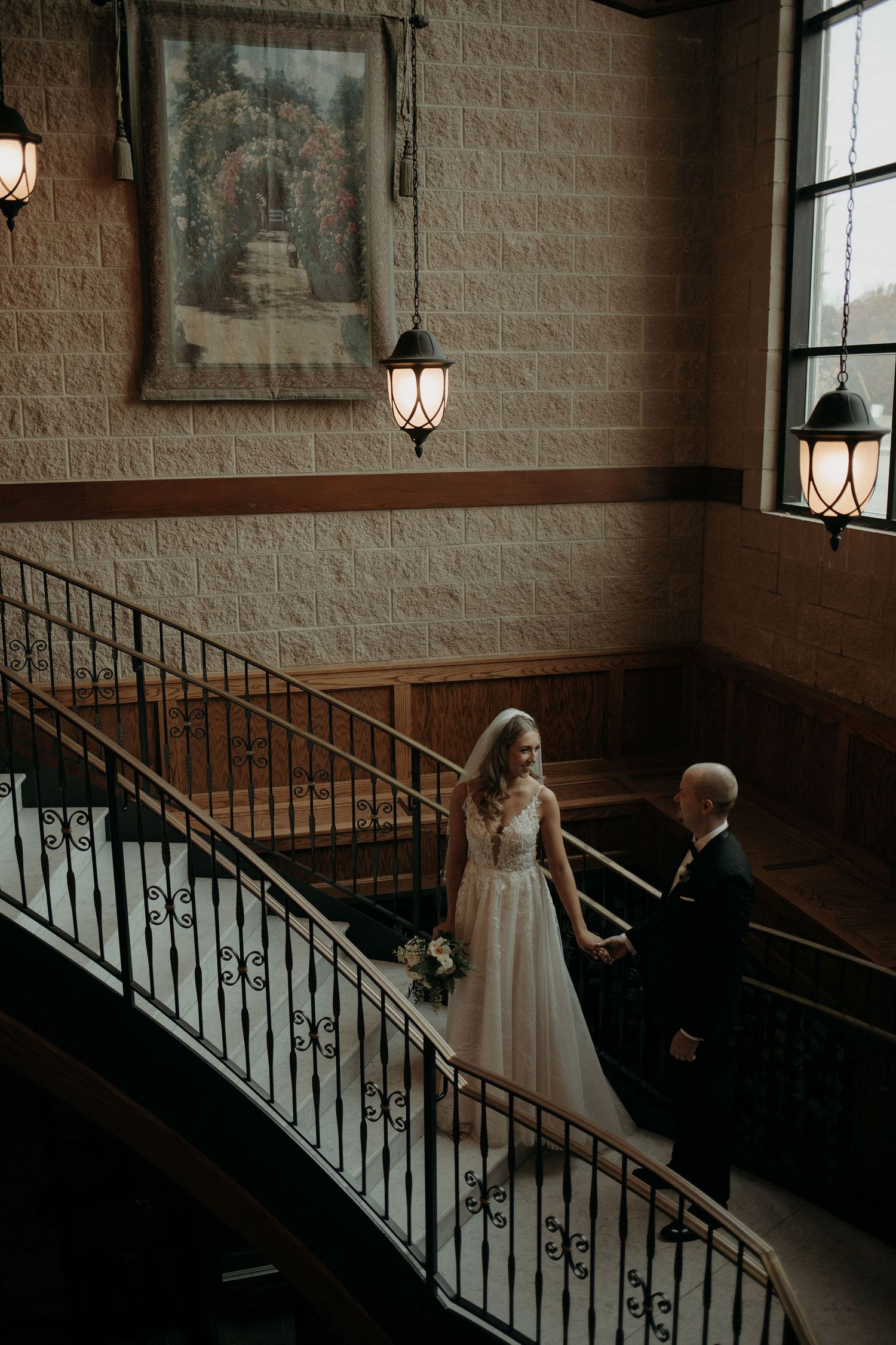 A couple descends an elegant curved staircase with wrought iron railings in a dimly lit interior with lantern sconces.