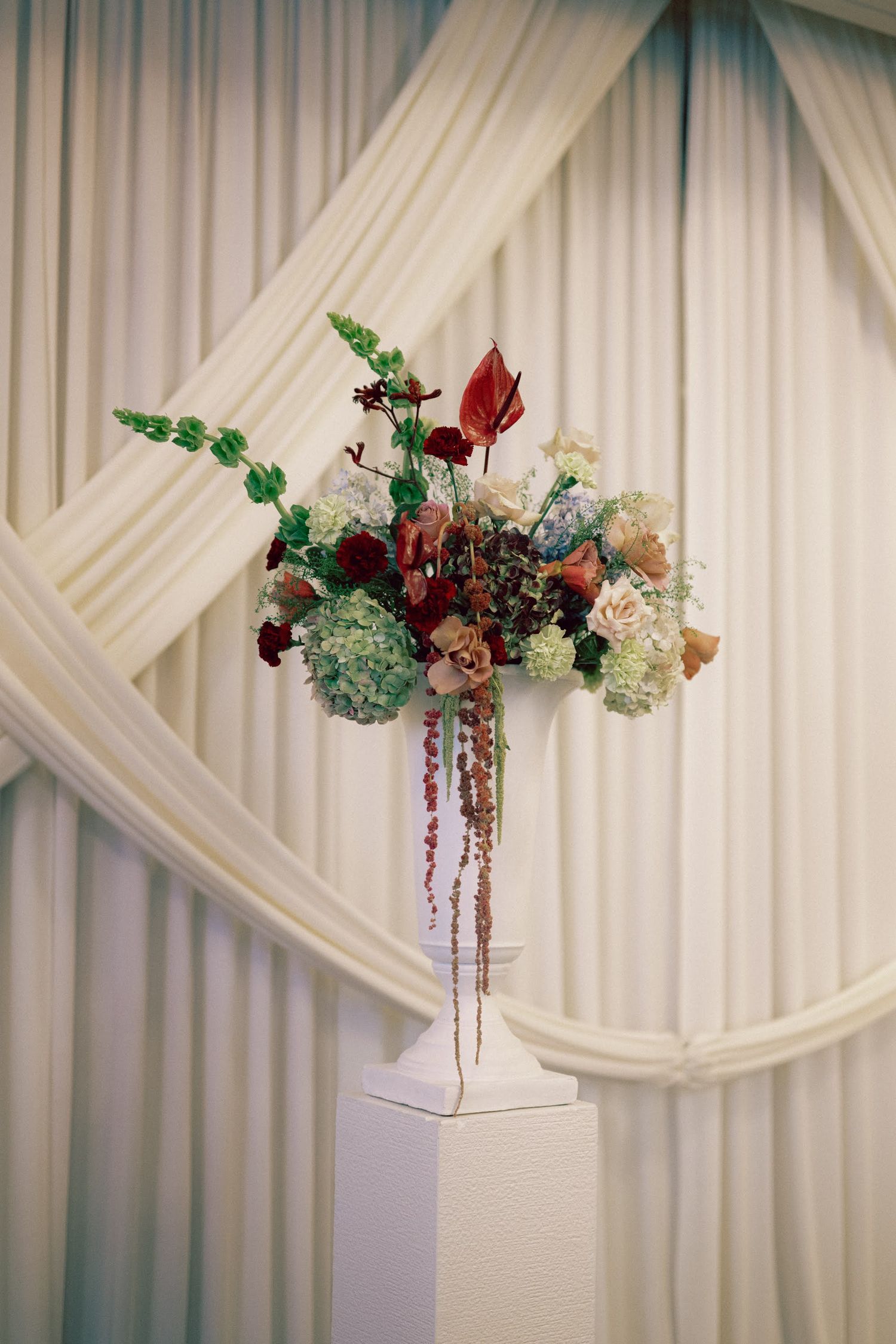 Elegant floral arrangement with hydrangeas, red anthurium, and greenery displayed on white pedestal against draped fabric backdrop.