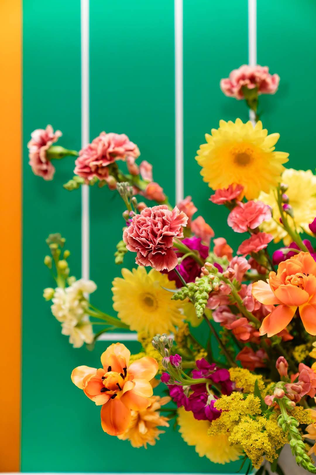 Vibrant bouquet of yellow gerberas, pink carnations, and orange flowers against a bright green wall.