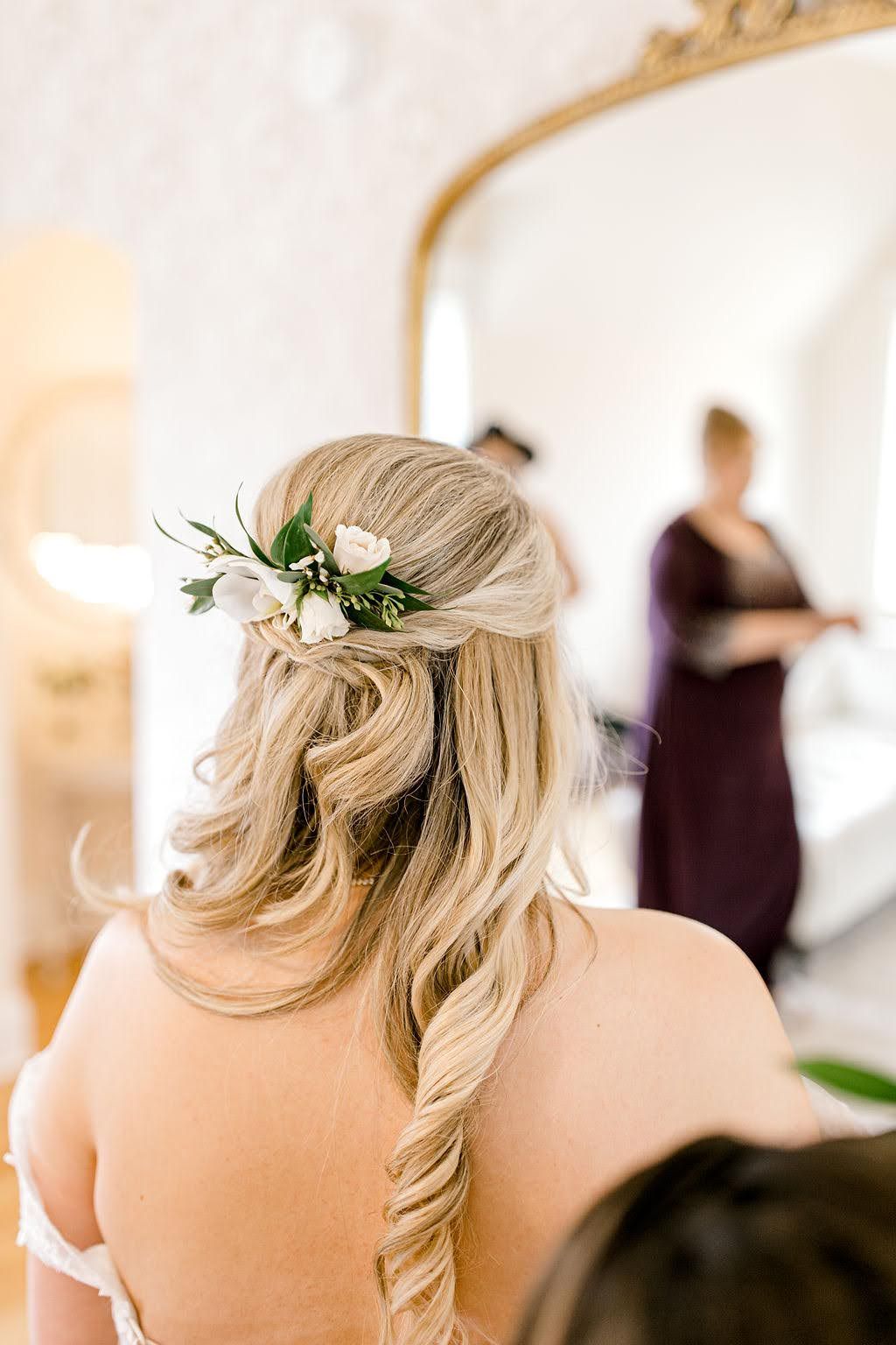 Bride with half-up hairstyle adorned with white flowers and greenery, reflected in an ornate gold mirror.