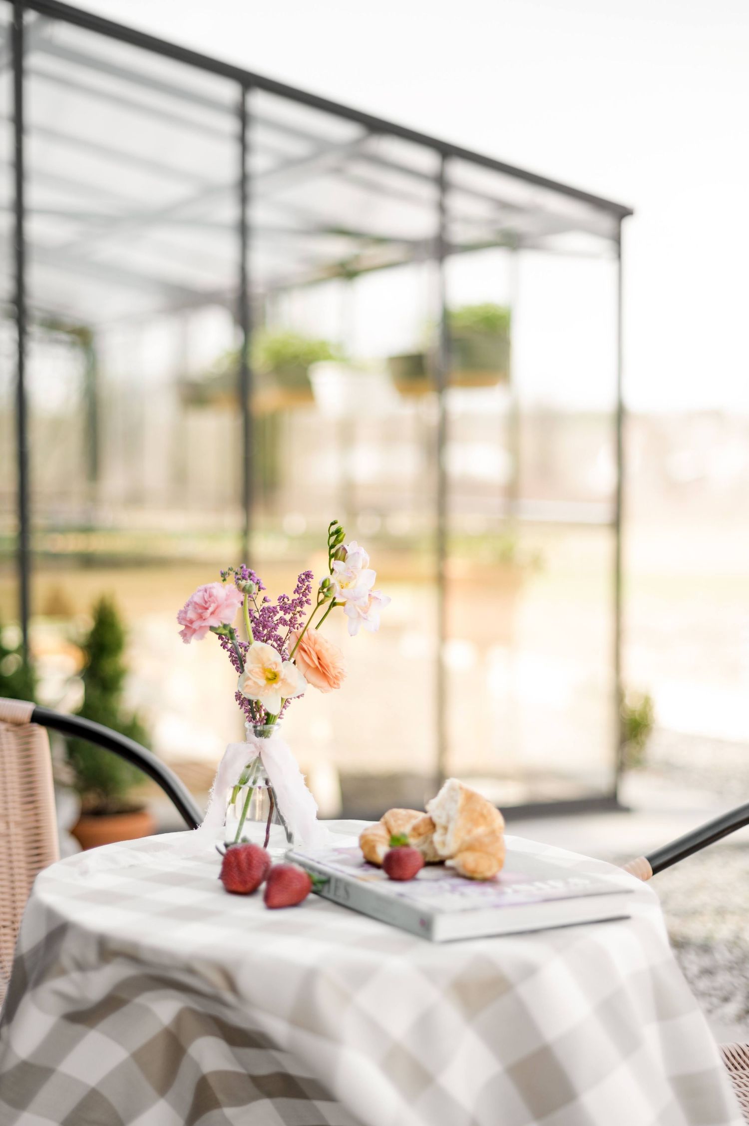 A romantic table setting with flowers in a vase, fresh strawberries, and croissants inside a bright greenhouse.