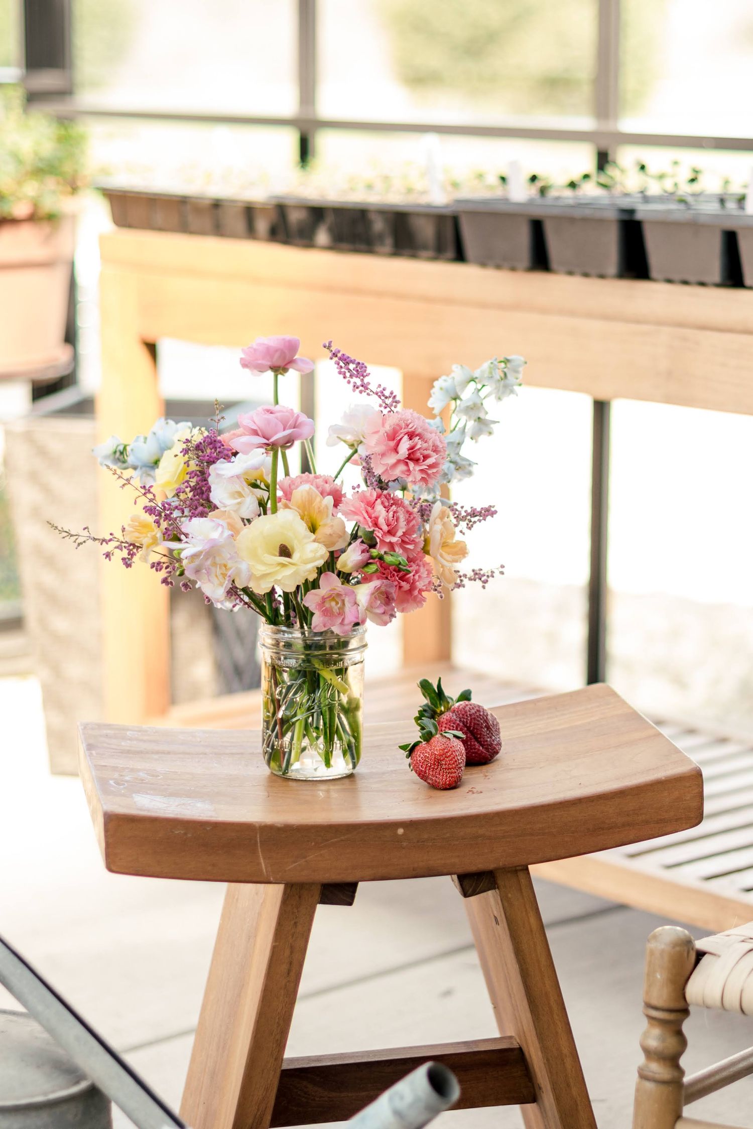 Fresh garden flowers arranged in a glass mason jar with strawberries on a rustic wooden stool by a window.