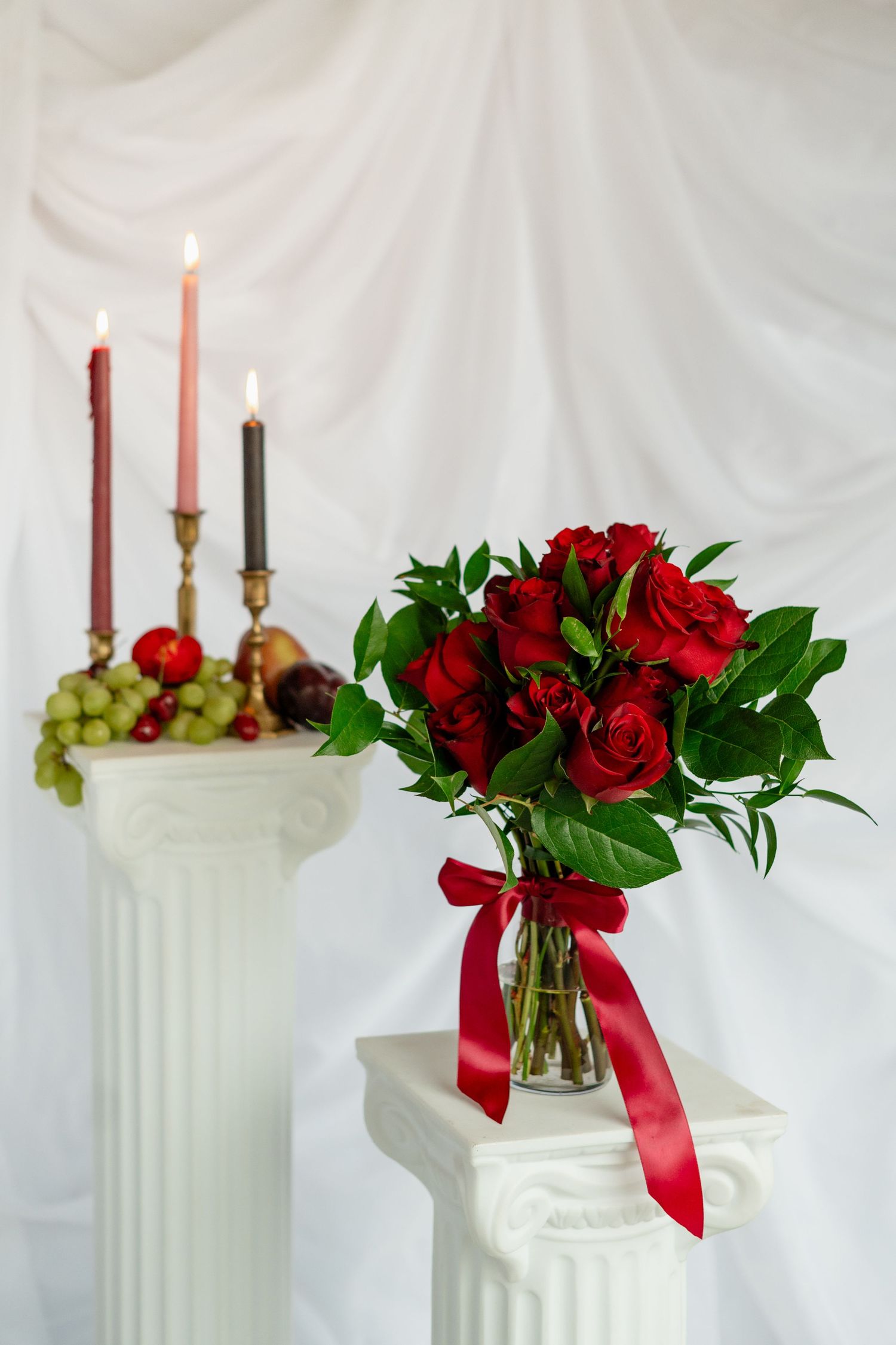Elegant white column displays with red roses, candles, and green grapes create a romantic classical setting against white backdrop.