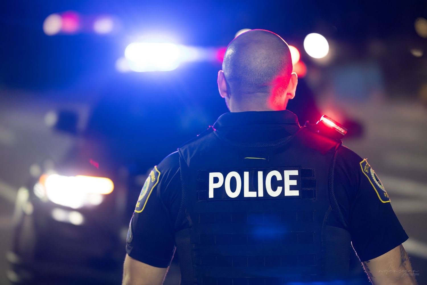 Police officer in uniform stands at night with blue and red emergency lights illuminating the background scene.