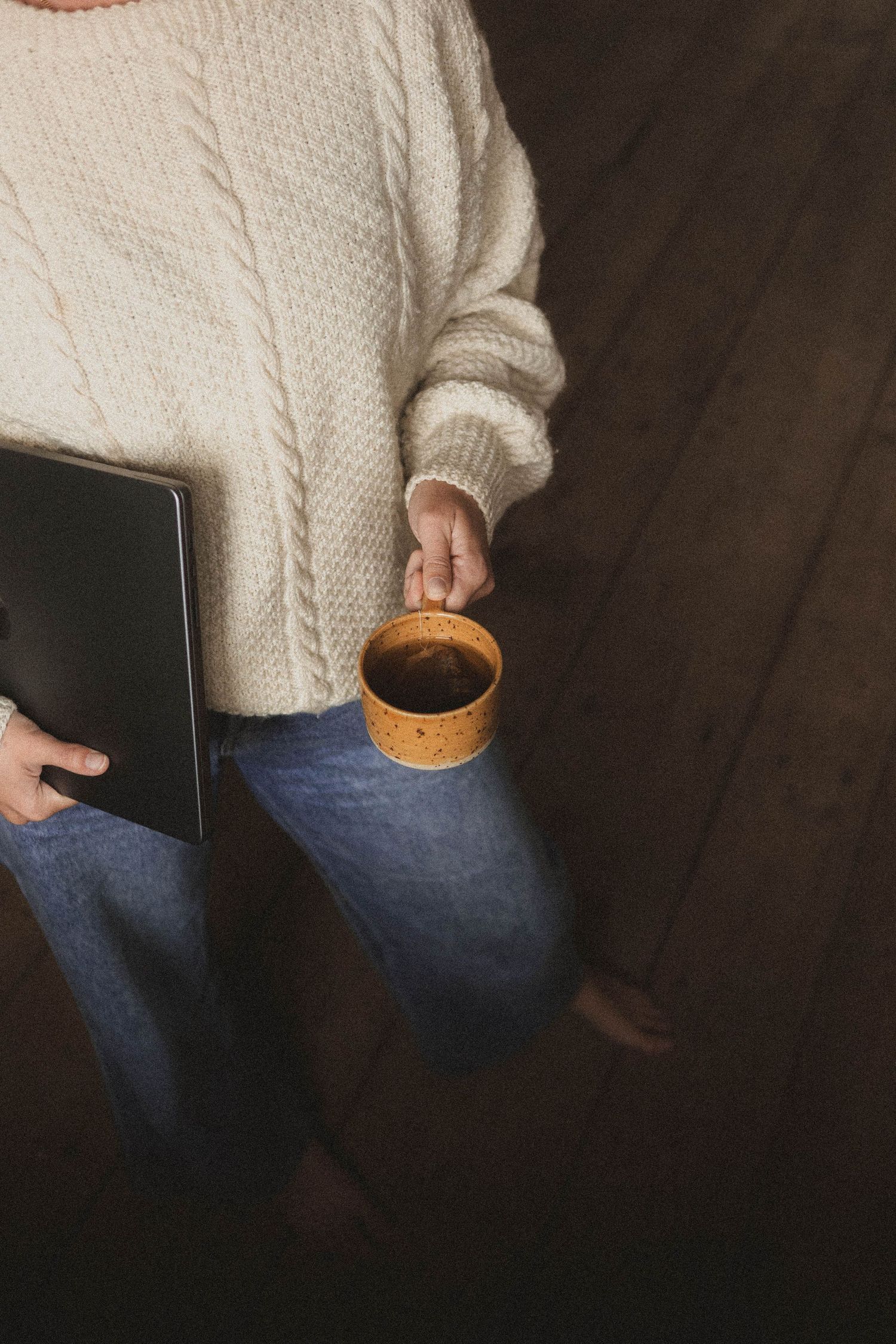Cropped view of someone in cream sweater and jeans holding a black laptop against wooden floor background.