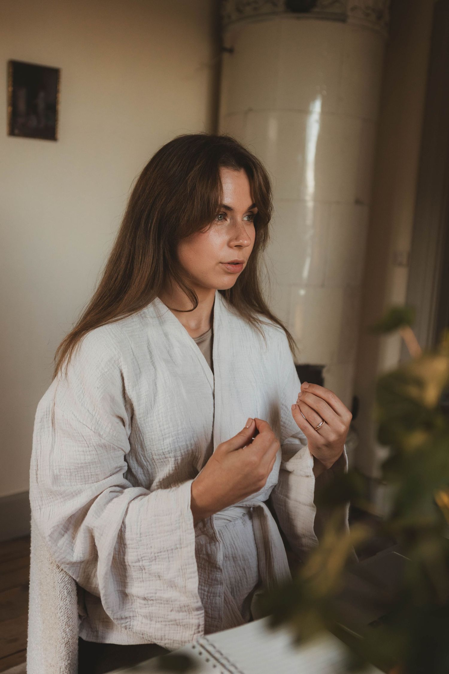 A person in a white linen shirt sits indoors while looking down in a contemplative pose with plants in the foreground.
