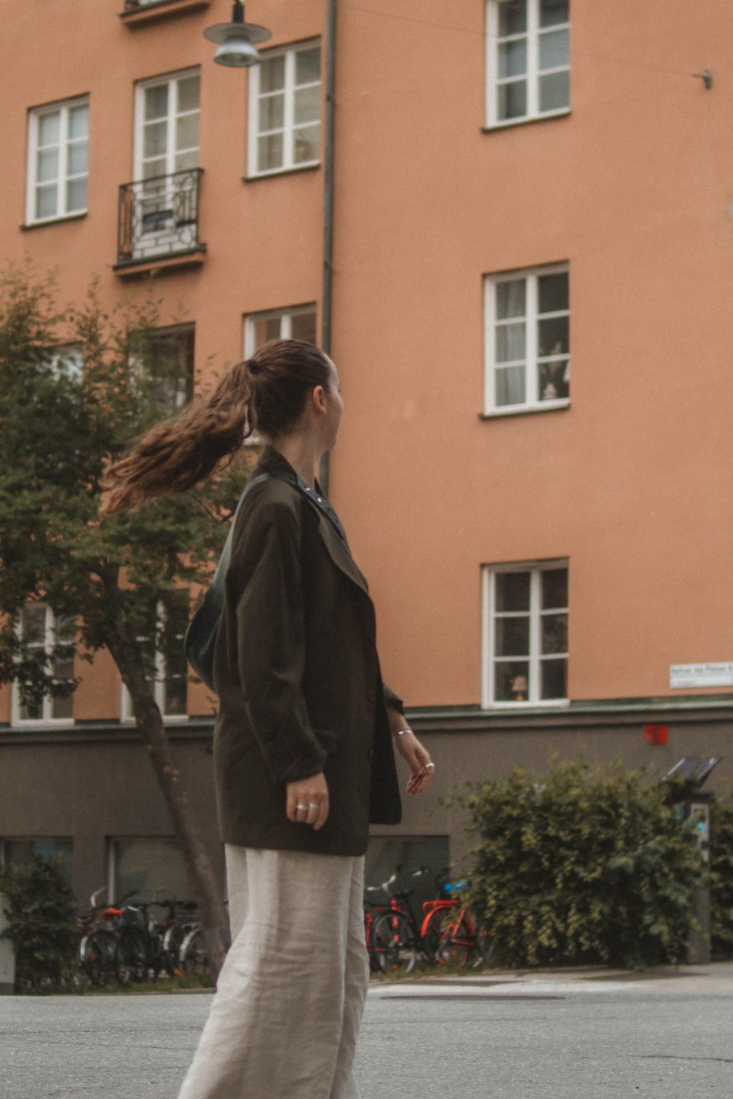 Someone with long hair wearing a dark blazer and light pants stands on a city street with a peach-colored apartment building behind.