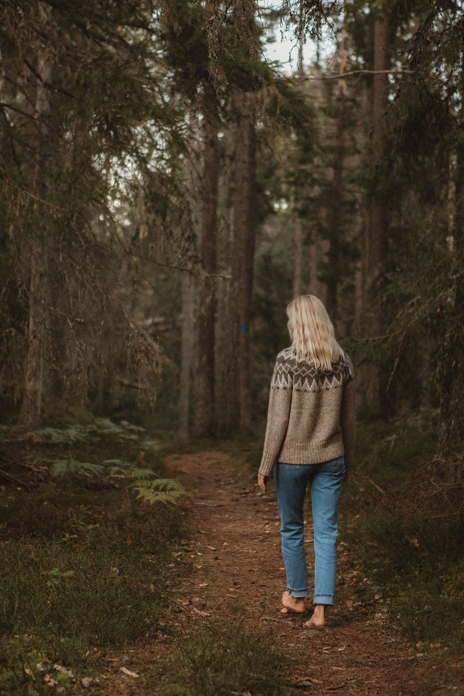 Person wearing patterned sweater walks along forest trail surrounded by pine trees.