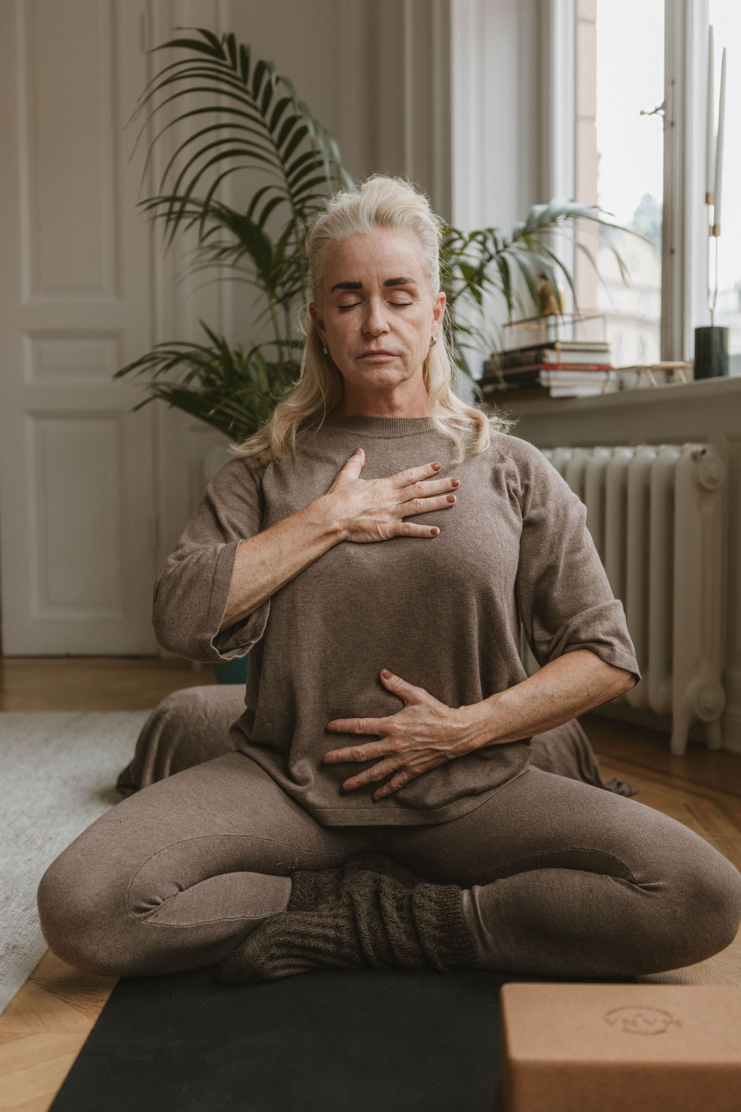 Person practices mindful breathing in a lotus pose on a yoga mat beside houseplants.