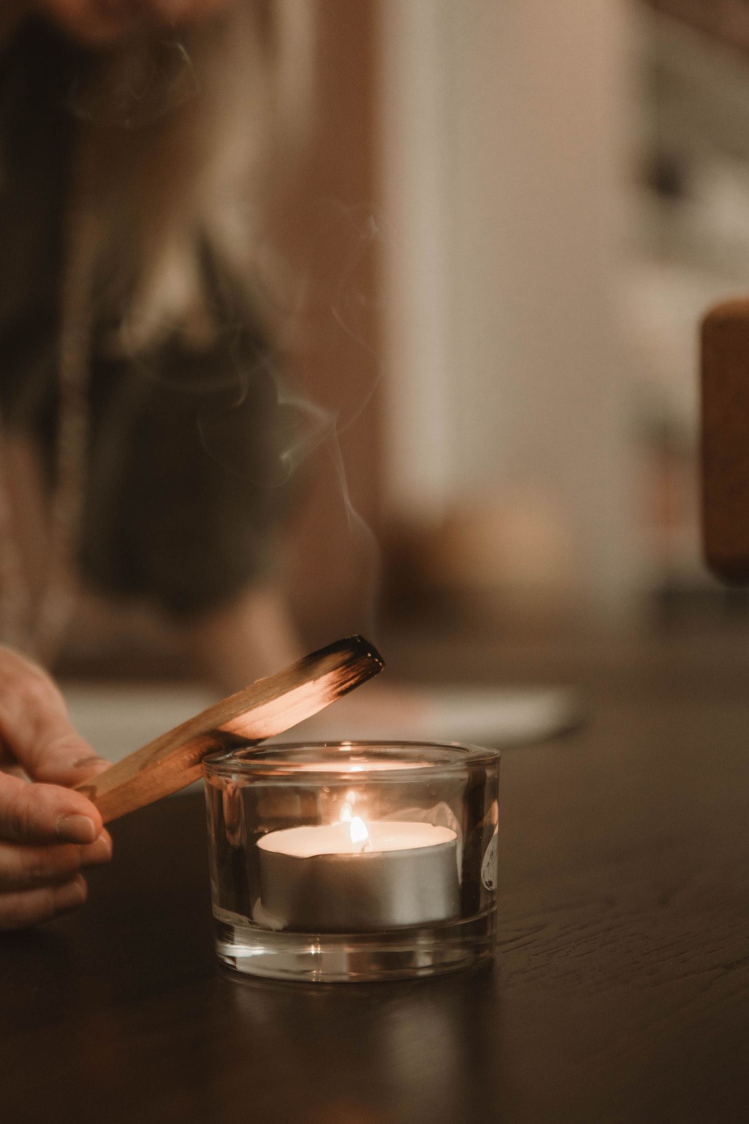Hands lighting a wooden stick from glowing tea light candle in glass holder on dark wooden surface.
