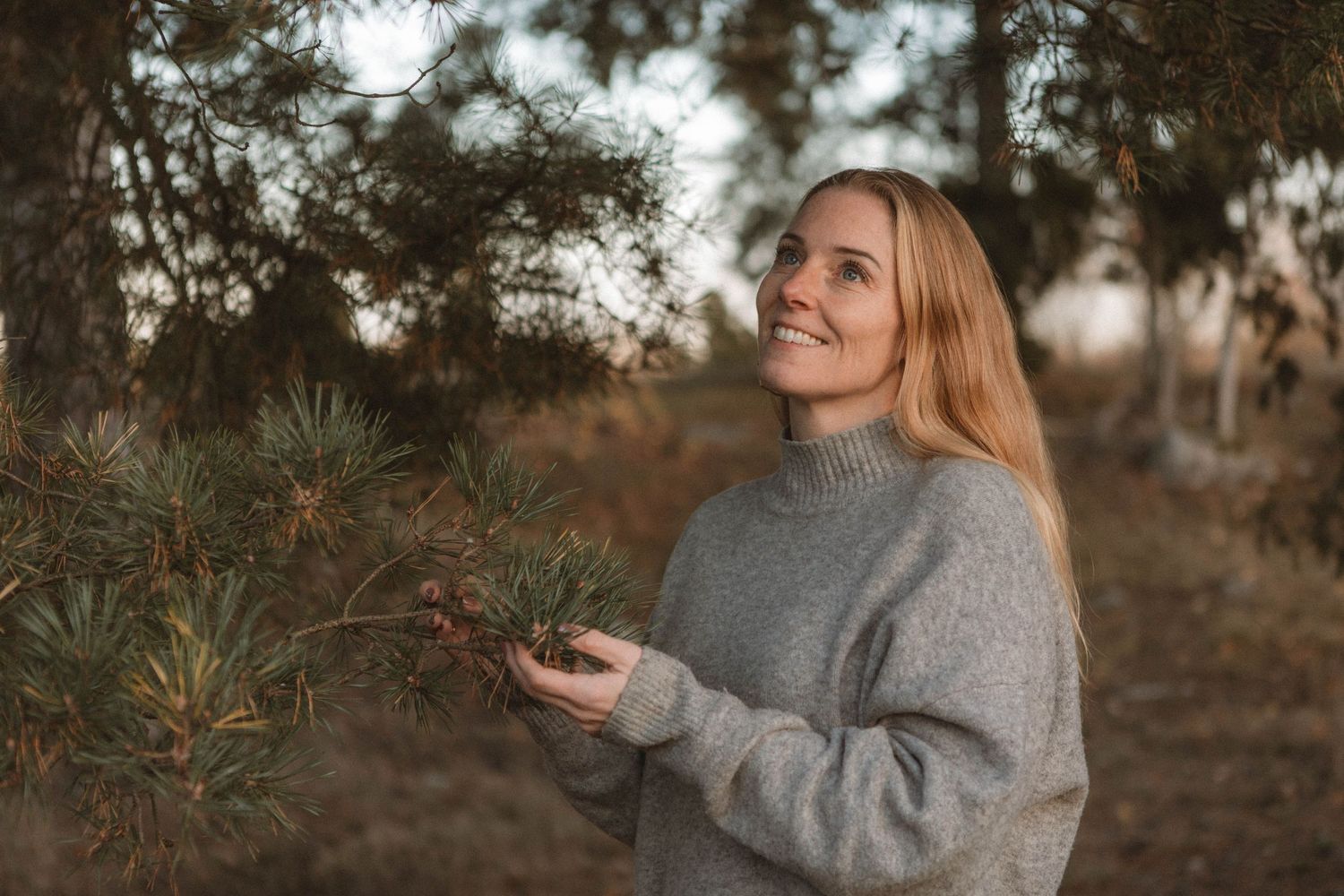 Someone in a cozy gray sweater stands in a pine forest during golden hour with a warm natural smile.