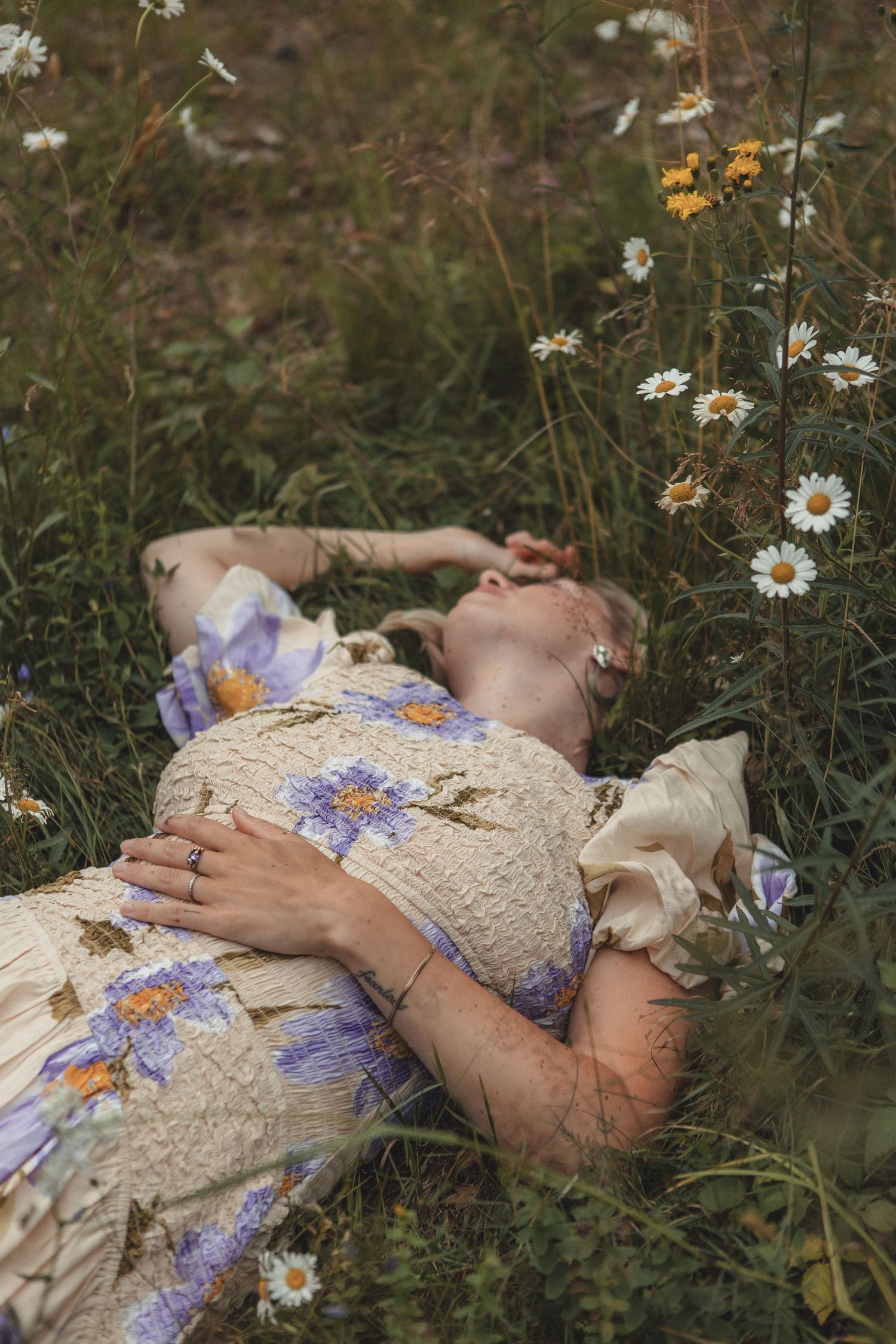 A person rests peacefully among wildflowers wearing a cream dress with purple floral pattern.