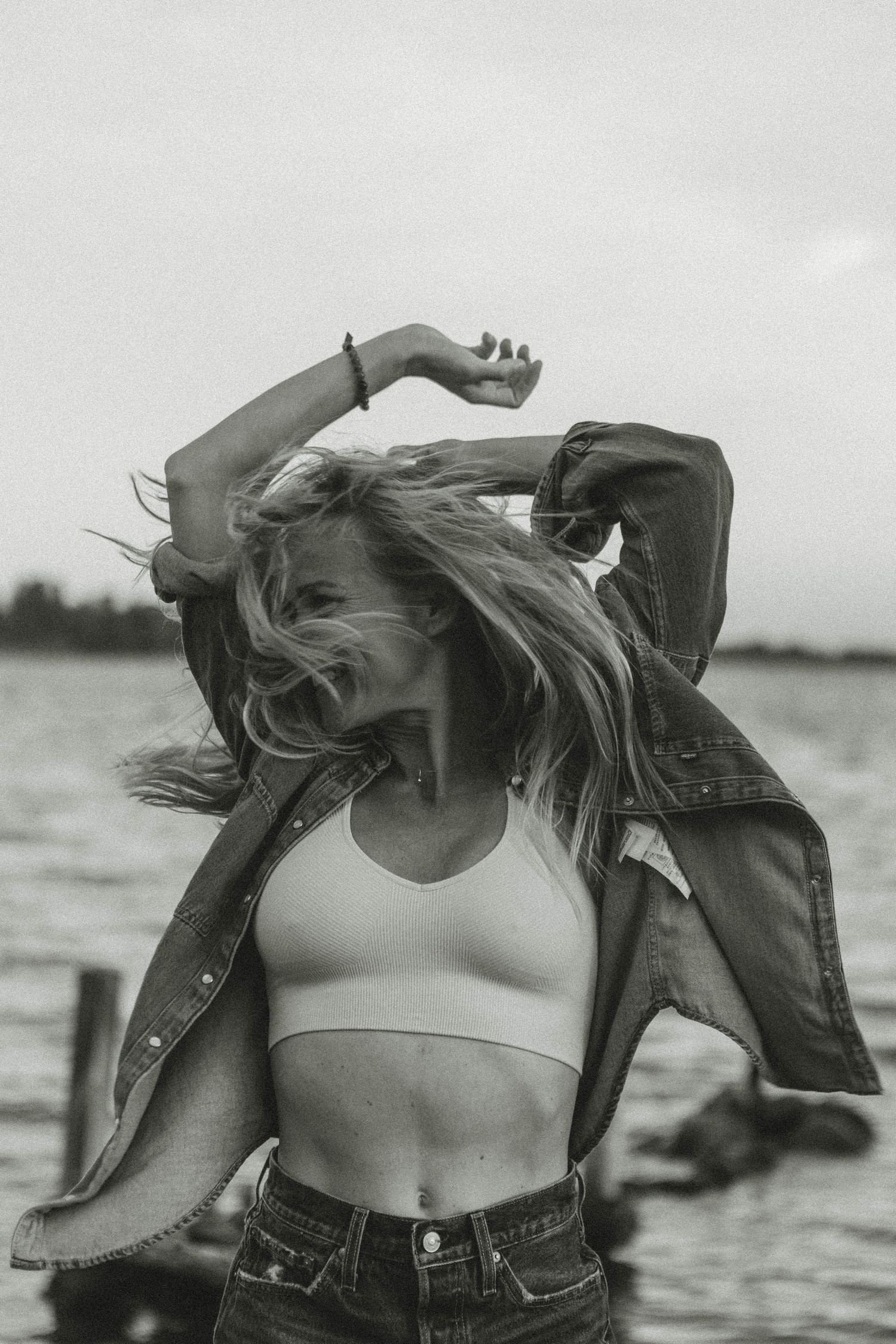 Person in crop top and denim jacket dances with wind-blown hair by water.