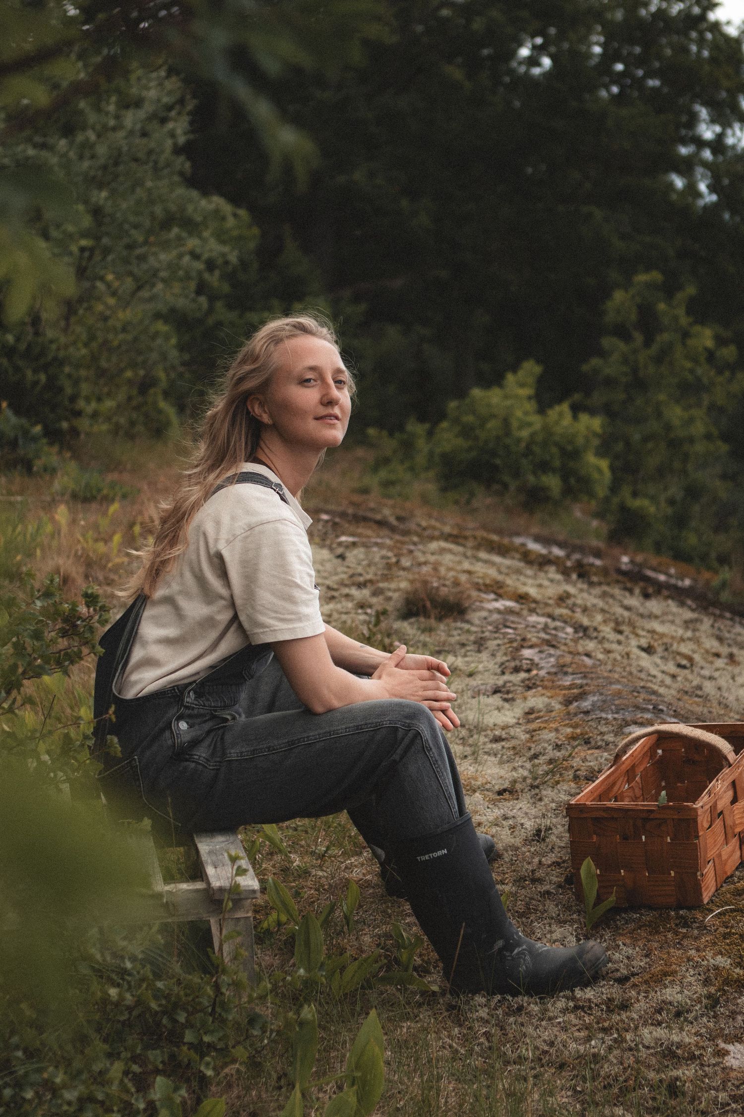 Person in overalls sits on hillside near wooden basket surrounded by natural landscape.
