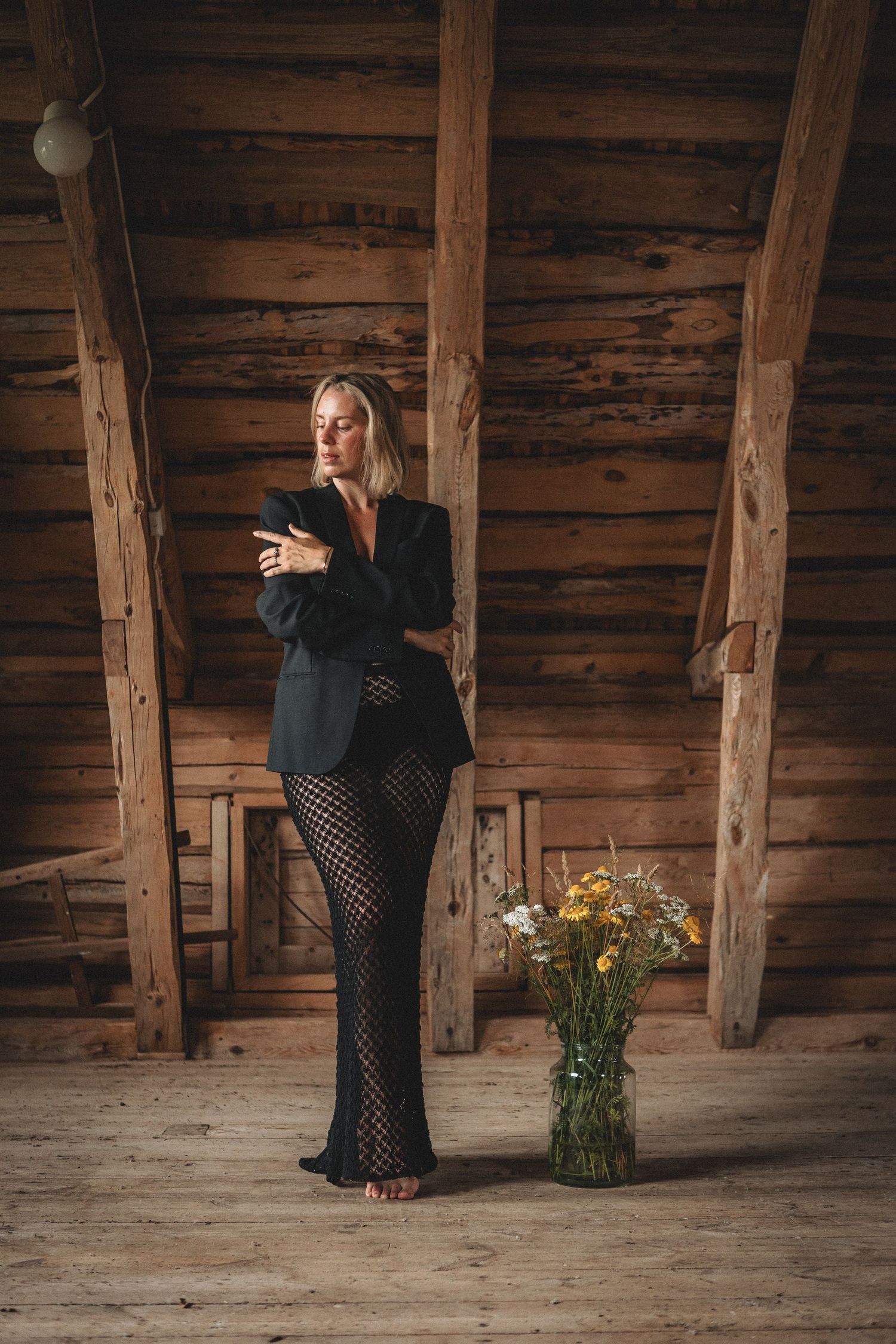 Person in elegant black outfit stands in rustic wooden barn interior with vase of flowers.