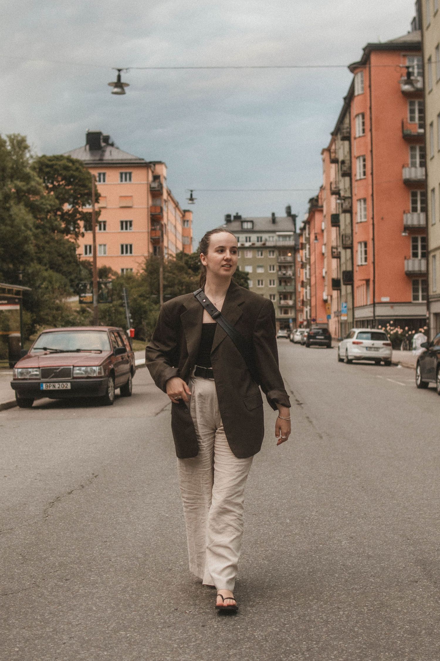 Someone in a dark blazer and light pants walks down a street lined with vintage-style apartment buildings in a city setting.