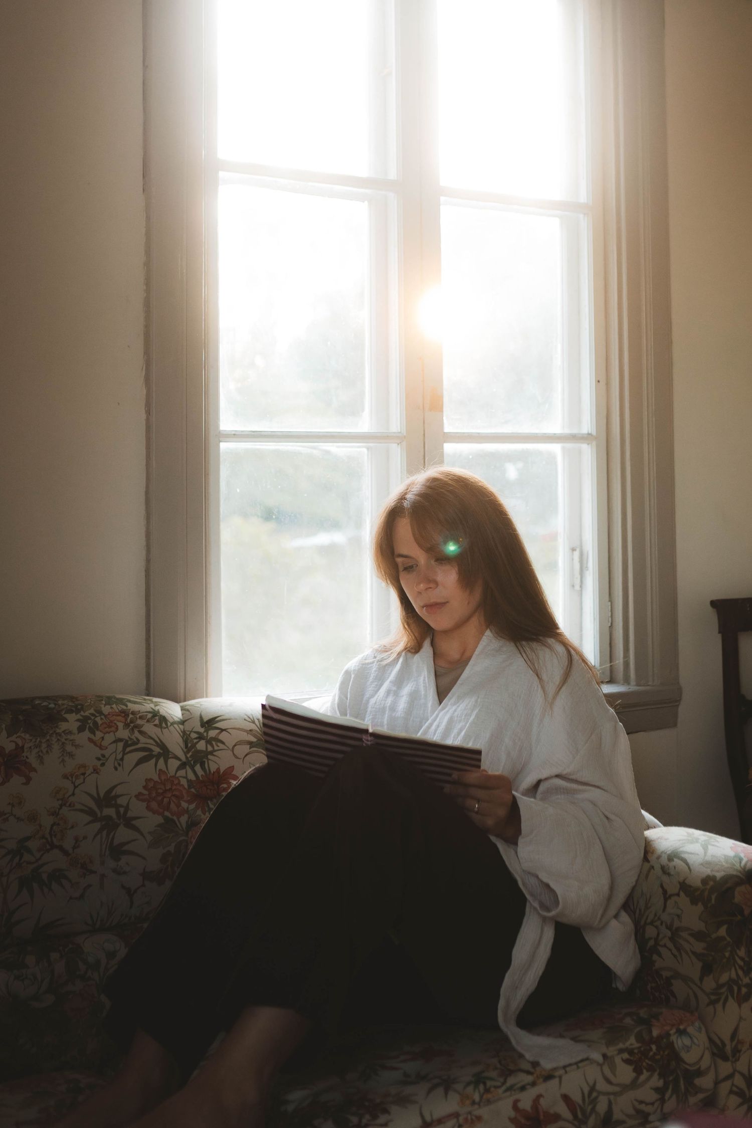 A person in a white bathrobe lounges on a floral couch near a sunlit window in a moody interior setting.