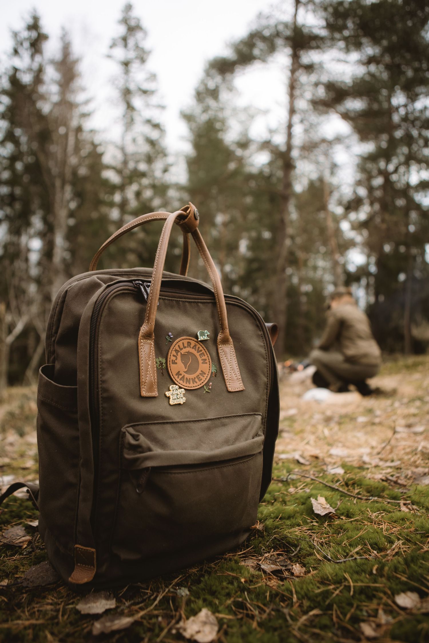 Dark green Fjallraven Kanken backpack with leather handles sitting on mossy forest ground with hiker in background.