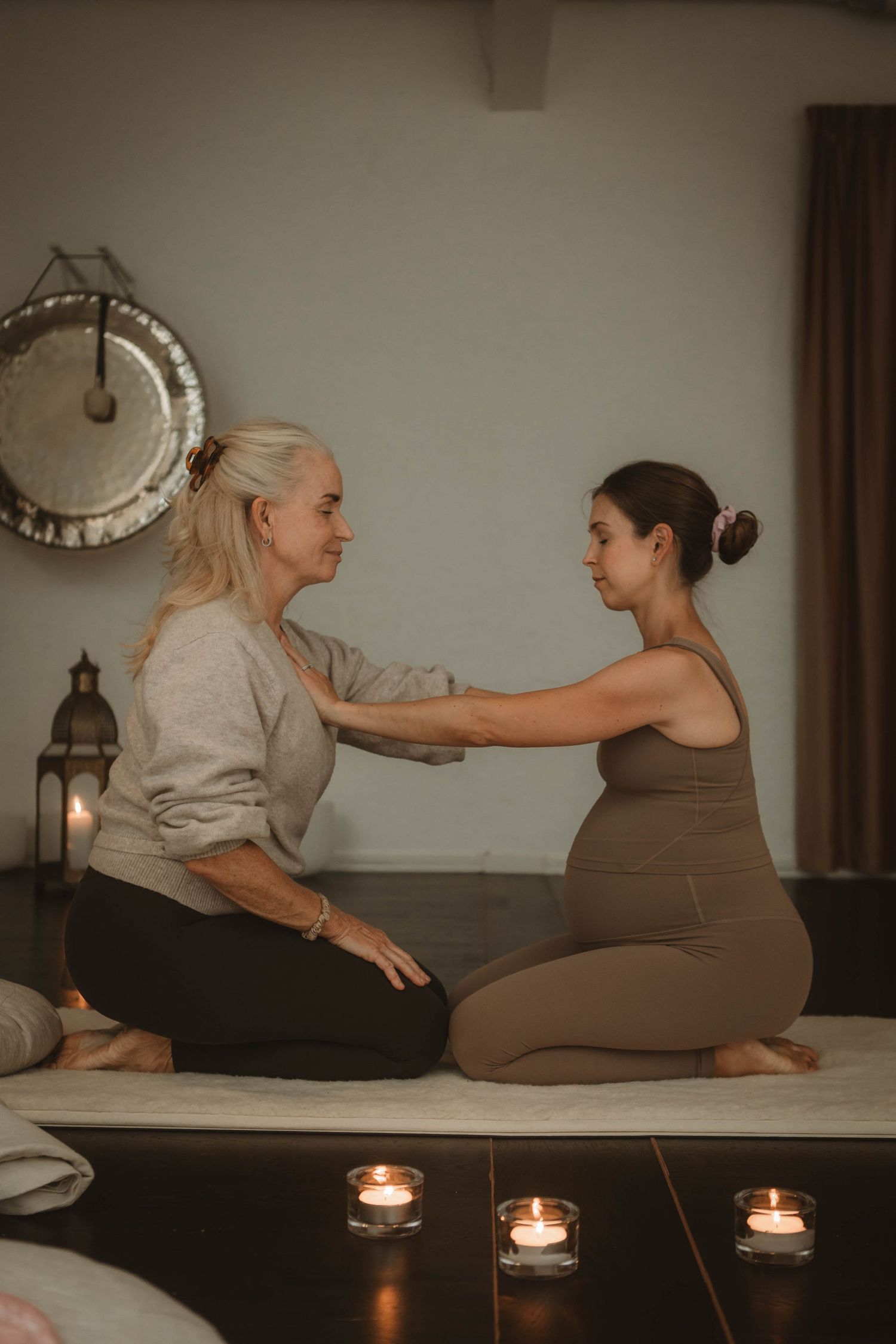 Two individuals sit on a mat in a calming wellness setting with lit candles and a decorative clock on the wall.