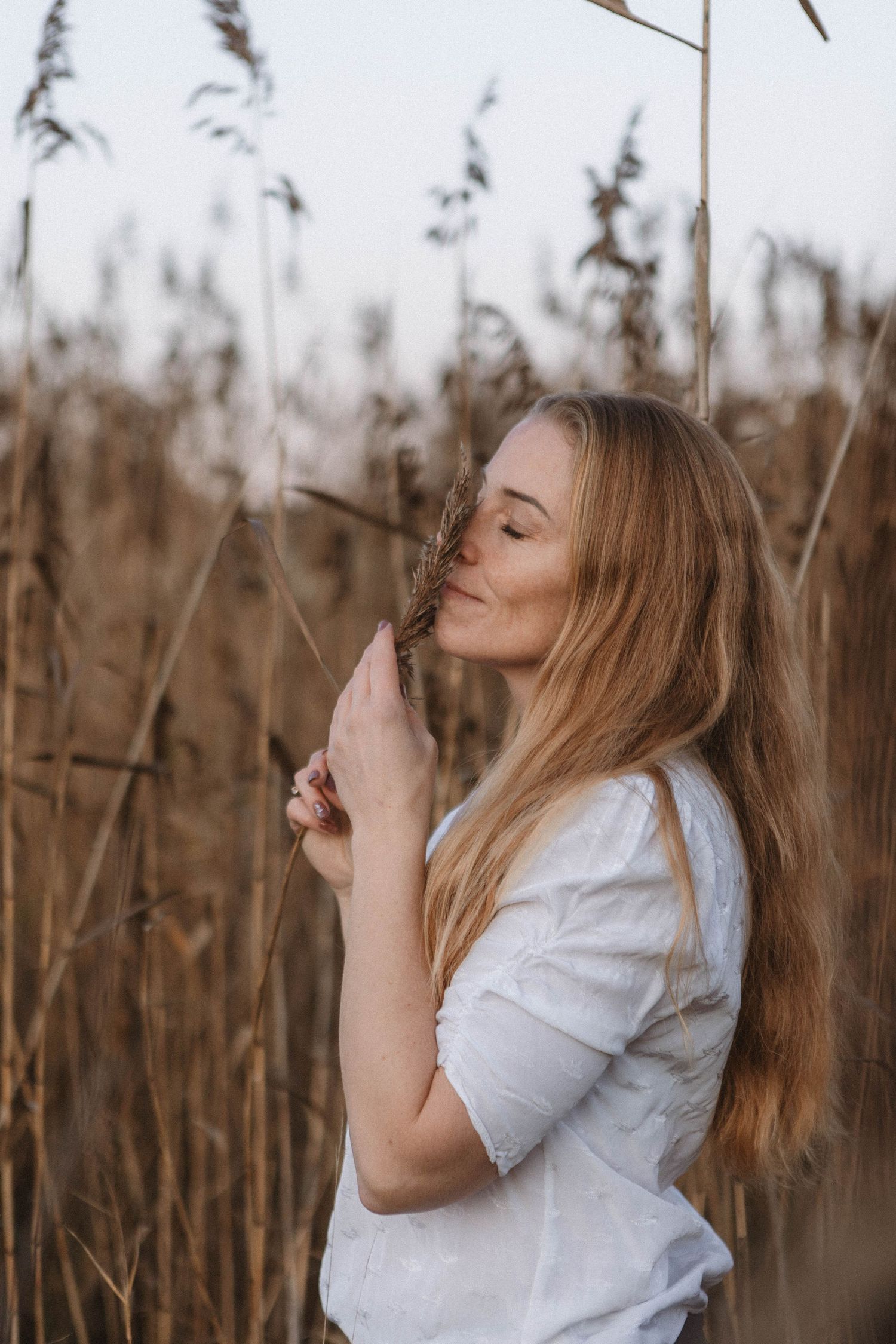 A woman in a white shirt stands serenely with her eyes closed among tall reed grass in a field at sunset.