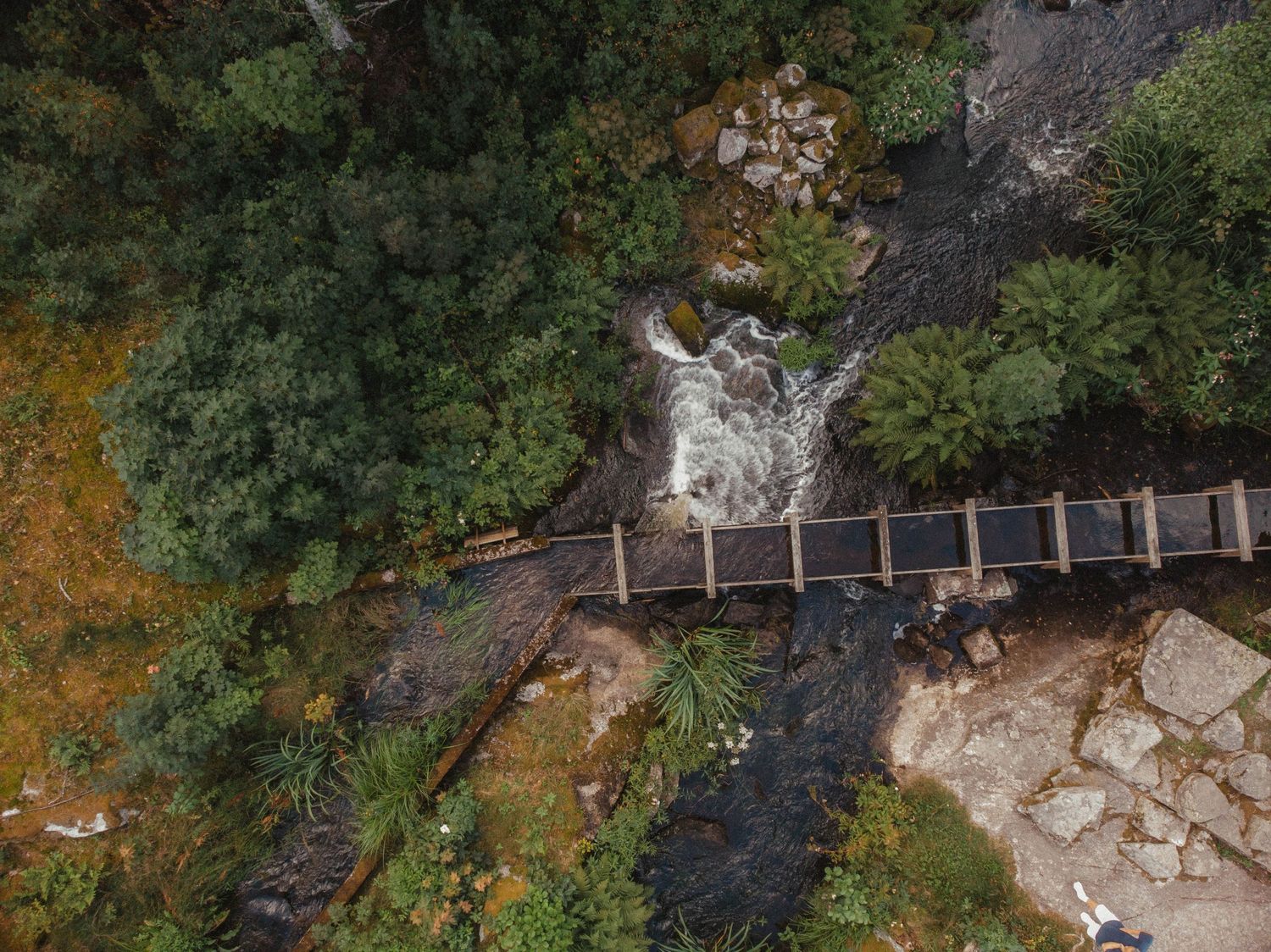 Aerial view of a wooden footbridge crossing over rushing water and rocks.