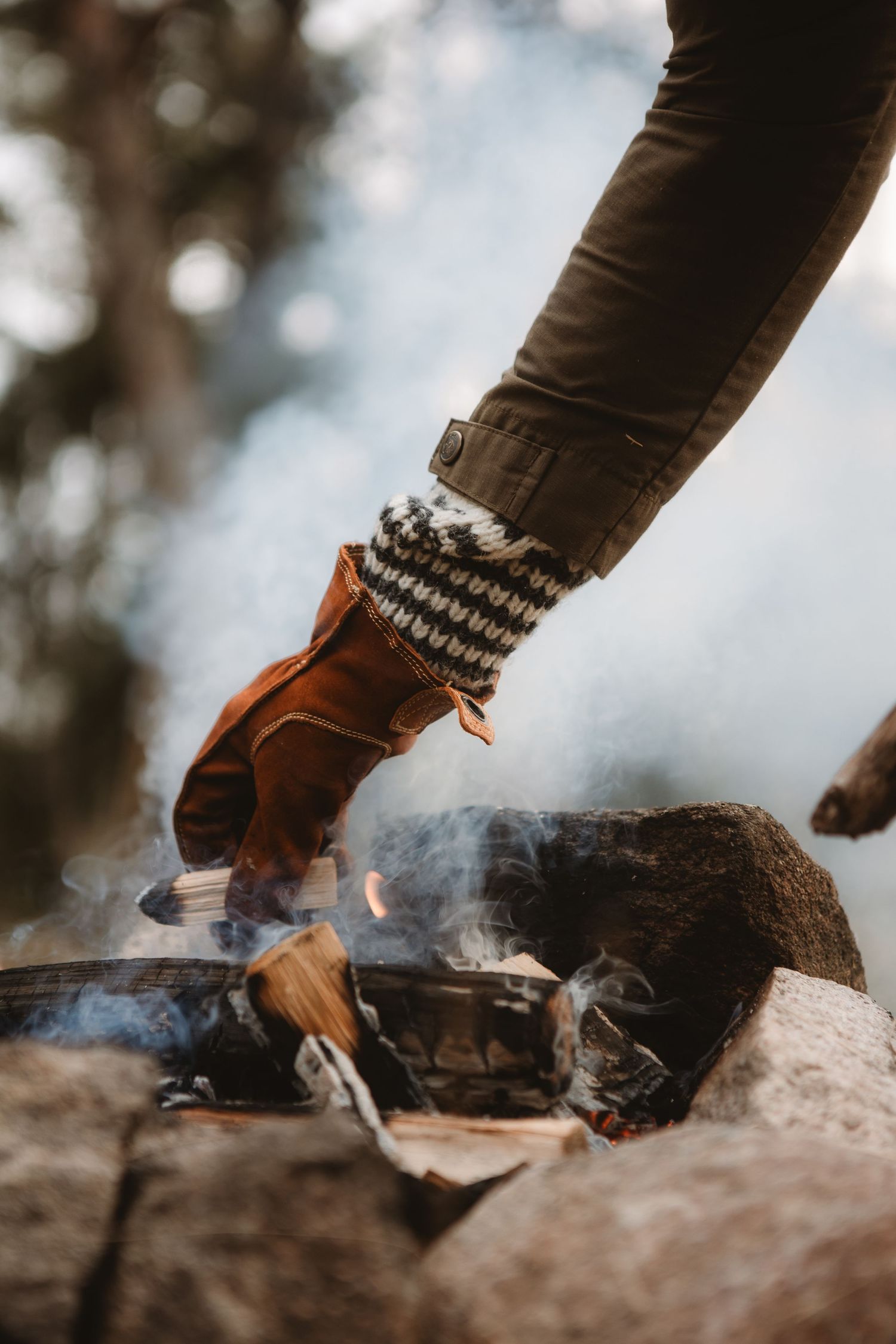 Person wearing knit gloves and leather mittens tending a campfire with burning logs and smoke outdoors.