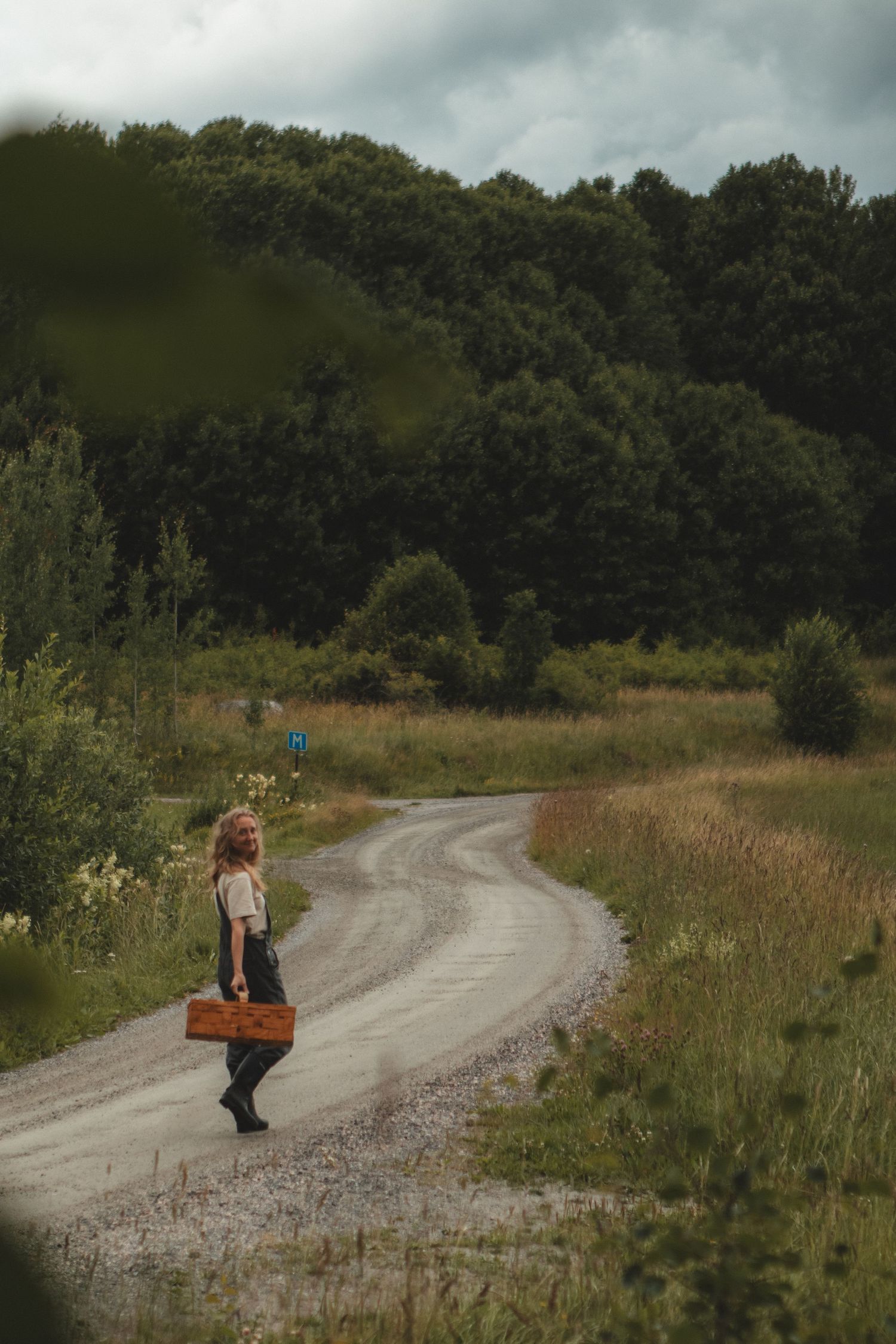 Person with leather bag stands on winding country dirt road surrounded by meadows.
